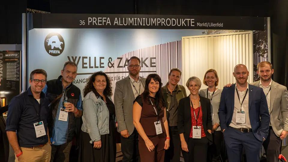 Am PREFA Messestand bei der Architect@Work in der Wiener Stadthalle präsentieren sich Fachexperten von PREFA aus den Bereichen Architekturberatung, Marketing und Vertrieb gemeinsam. Vor der PREFA Leinwand, die moderne Wellen- und Zackenprofile darstellt und die vielfältigen Gestaltungsmöglichkeiten zeigt, steht die Gruppe. PREFA Aluminium überzeugt durch Langlebigkeit, Leichtigkeit und nachhaltige Lösungen für anspruchsvolle Architektur.