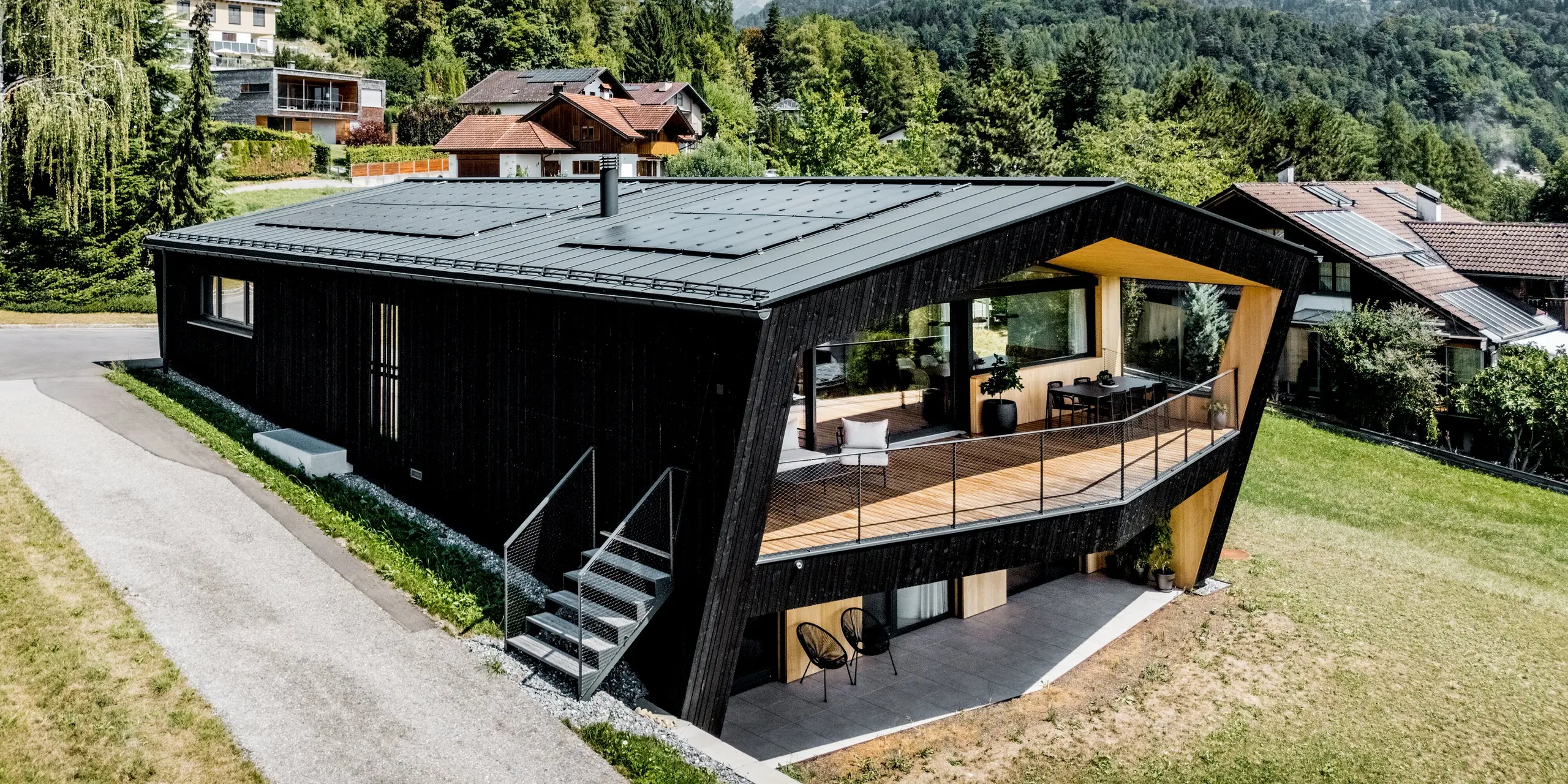 Beeindruckende Schrägansicht des modernen Einfamilienhauses in Röthis: Das PREFALZ Satteldach in P.10 Anthrazit mit integrierten Solarmodulen PREFALZ krönt die markante, dunkle Holzfassade. Besonders ins Auge fällt der großzügige Balkon mit warmer Holzverkleidung und filigranem Geländer. Die langlebige, leichte und witterungsbeständige Aluminiumdeckung verbindet ausdrucksstarke Architektur mit zuverlässigem Schutz und nachhaltiger Energiegewinnung.