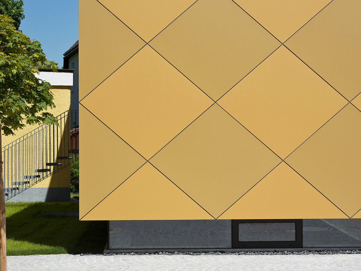 Detailaufnahme der FAssade des Musikvereins in Zwettl. Im unteren Bereich des Gebäudes wurden Fenster installiert, die für eine bessere Lichtversorgung im Innenbereich sorgen. Die Außenhülle wurde mit goldfarbenen Aluminium Verbundplatten von PREFA verkleidet.