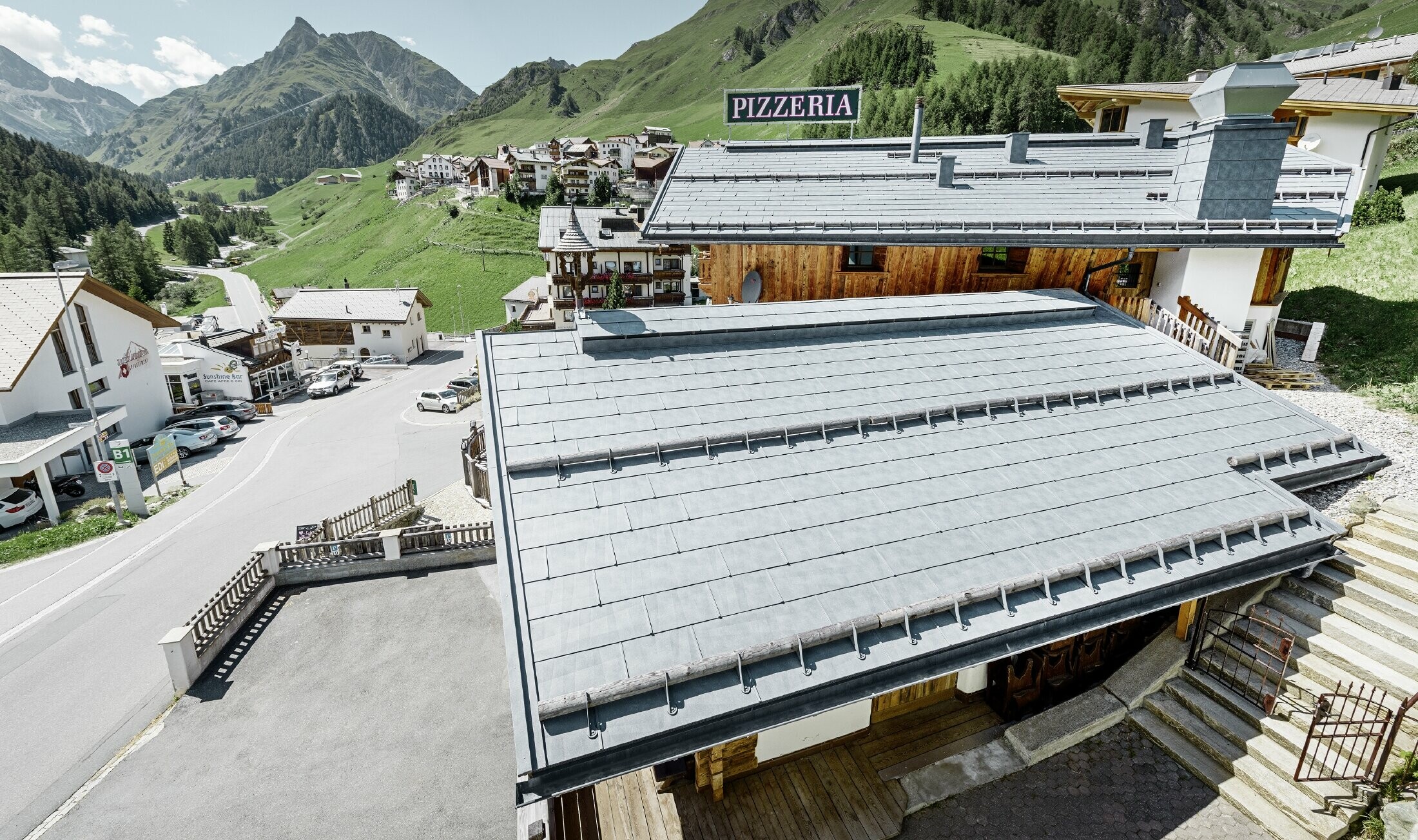 Restaurant Almrausch mit Bergen im Hintergrund und einem PREFA Dach mit dem Dachpaneel FX.12 in steingrau und Gebirgsschneestützen