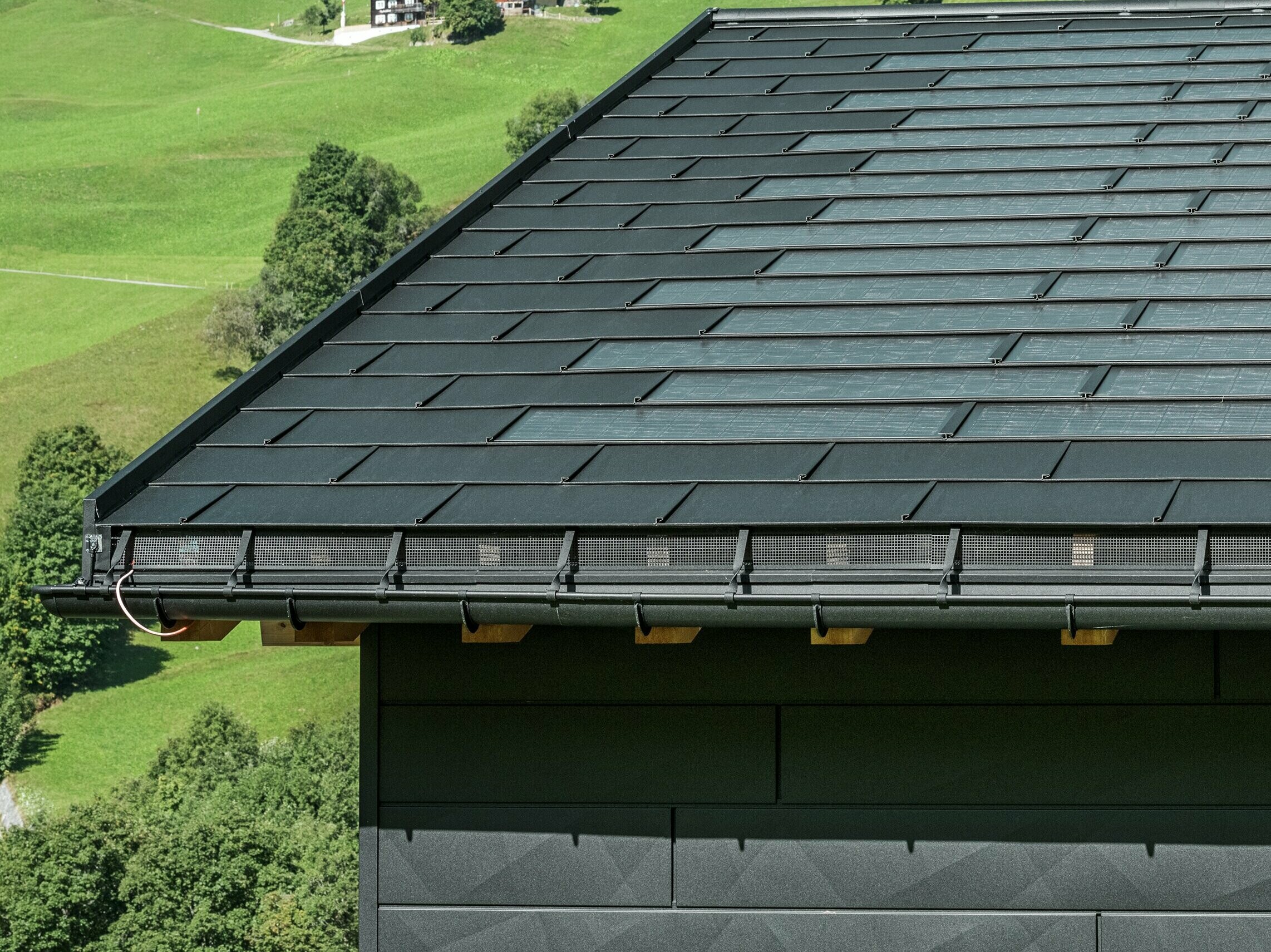 Nahaufnahme der Traufe eines modernen Einfamilienhauses in der alpinen Umgebung von Ascharina in der Schweiz. Das Dach wurde mit der innovativen PREFA Solardachplatte und der traditionellen Dachplatte R.16, beide in P.10 Schwarz, eingedeckt. Für die zuverlässige Dachentwässerung sorgen die PREFA Hängerinne und das PREFA Ablaufrohr. Am unteren Bildrand sind die unverwechselbaren PREFA Siding.X Fassadenpaneele zu sehen. Die klare Linienführung der Aluminiumprodukte unterstreicht das moderne Design inmitten einer malerischen Berglandschaft.