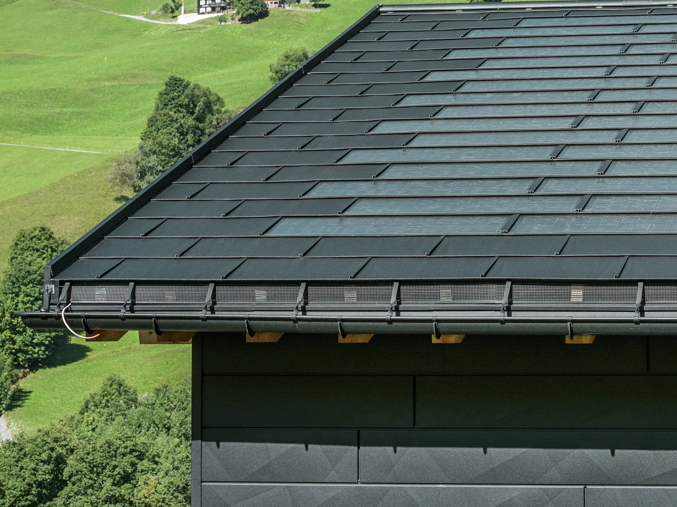 Nahaufnahme der Traufe eines modernen Einfamilienhauses in der alpinen Umgebung von Ascharina in der Schweiz. Das Dach wurde mit der innovativen PREFA Solardachplatte und der traditionellen Dachplatte R.16, beide in P.10 Schwarz, eingedeckt. Für die zuverlässige Dachentwässerung sorgen die PREFA Hängerinne und das PREFA Ablaufrohr. Am unteren Bildrand sind die unverwechselbaren PREFA Siding.X Fassadenpaneele zu sehen. Die klare Linienführung der Aluminiumprodukte unterstreicht das moderne Design inmitten einer malerischen Berglandschaft.
