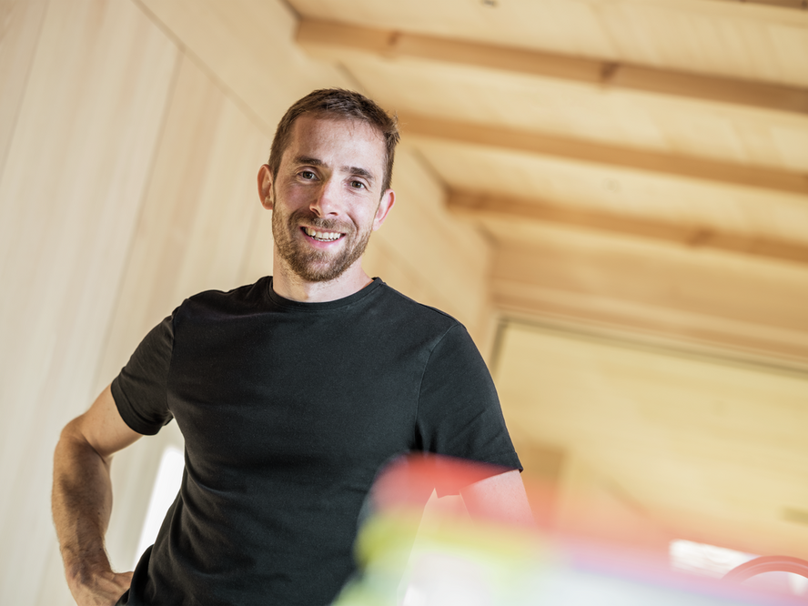 Porträt von Daniel Zimmermann, dem Bauherrn und Planer, in einem hellen Raum mit Holzverkleidung. Er lehnt sich lächelnd zurück und trägt ein schwarzes T-Shirt. 