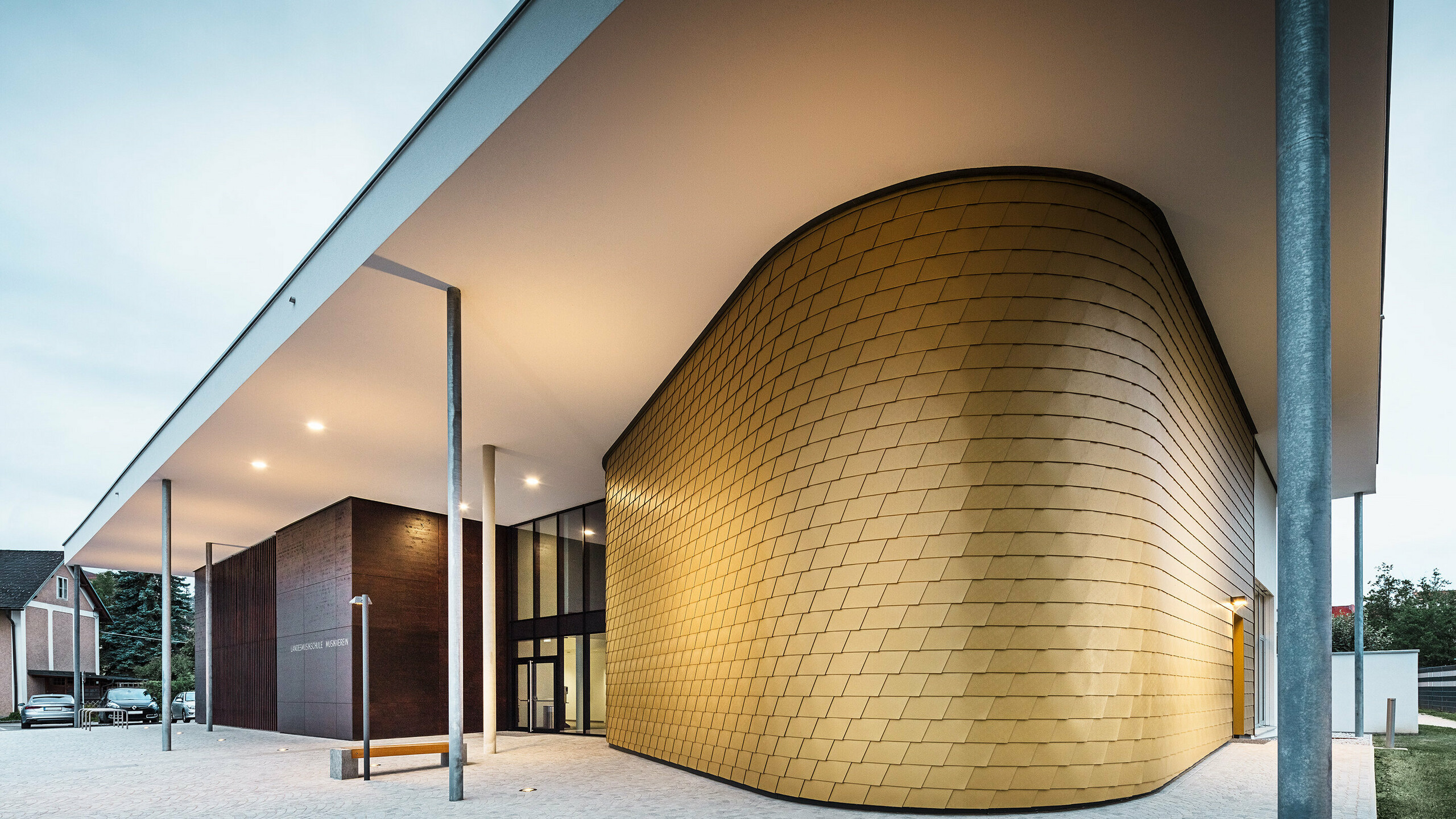 Modernes Gebäude mit markantem Eingangsbereich, dessen geschwungene Fassade mit goldfarbenen Aluminiumrauten verkleidet ist. Das Vordach wird von schlanken Stützen getragen und ist dezent beleuchtet. Neben dem goldenen Fassadenteil befindet sich ein dunkler Kubus mit der Aufschrift „Landesmusikschule Buchkirchen“. Der Vorplatz ist mit hellen Pflastersteinen gestaltet.