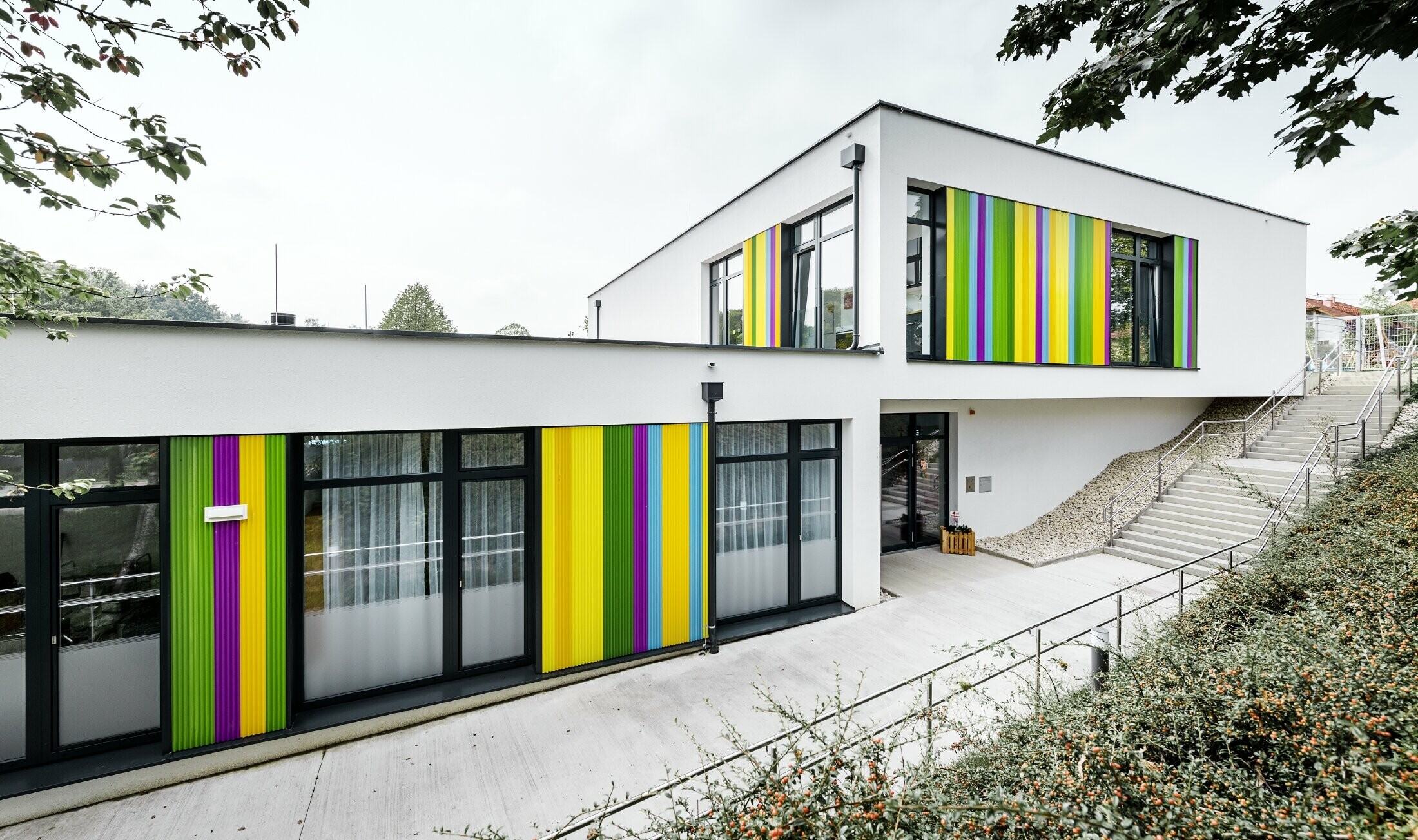 Bunte Fassadenelemente von PREFA für den Kindergarten in Hargelsberg. Das Gebäude hat ein Flachdach und große, bodentiefe Fenster.