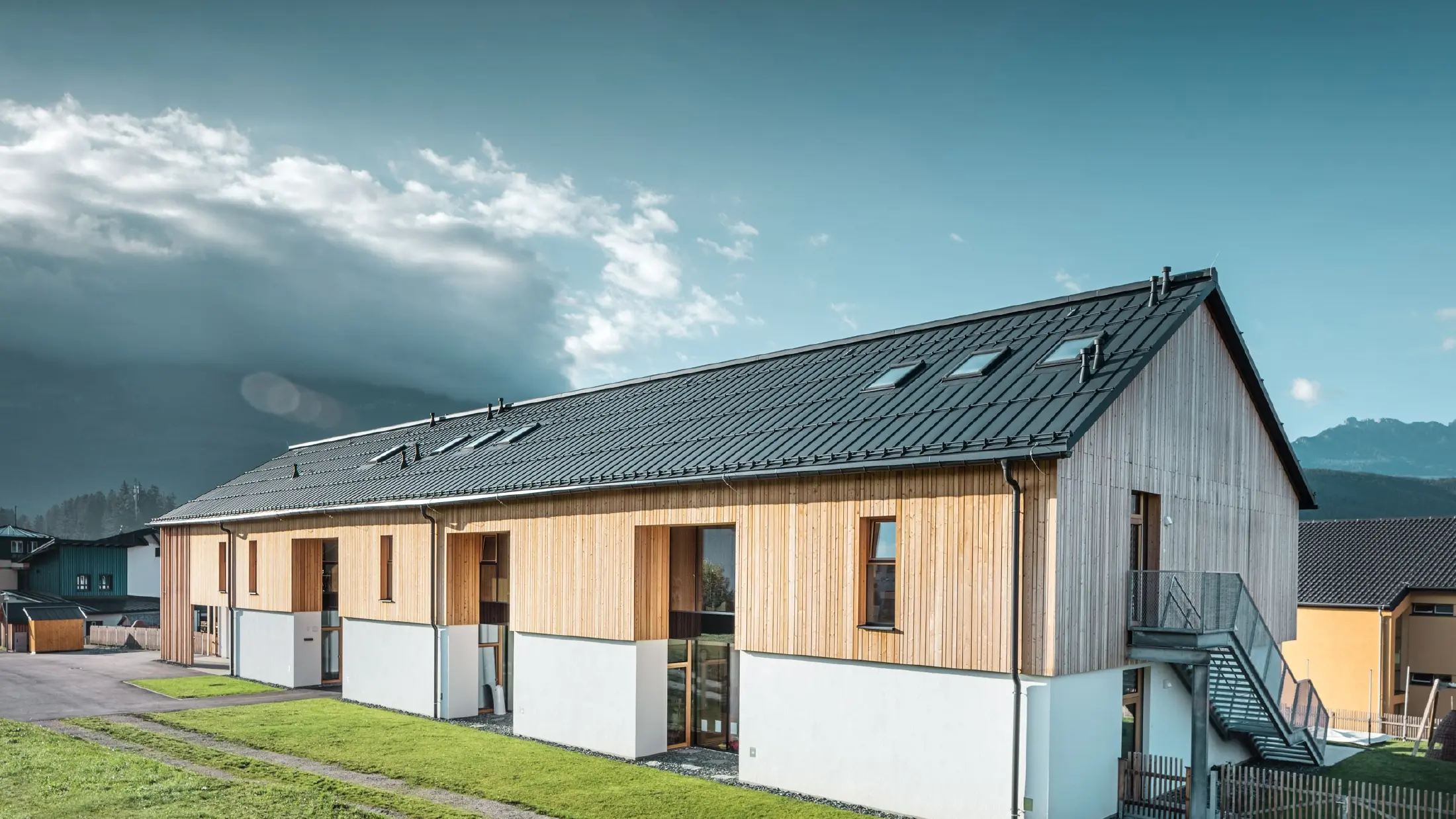 Modernes, langgestrecktes Gebäude mit Satteldach aus dunkelgrauem Aluminium und Fassade aus vertikaler Holzverschalung über weißem Sockel. Große Fensteröffnungen und eine Außentreppe aus Metall betonen die funktionale Architektur. Das Gebäude steht auf einer grünen Wiese mit umliegenden Häusern und Bergen im Hintergrund unter teils wolkigem Himmel.
