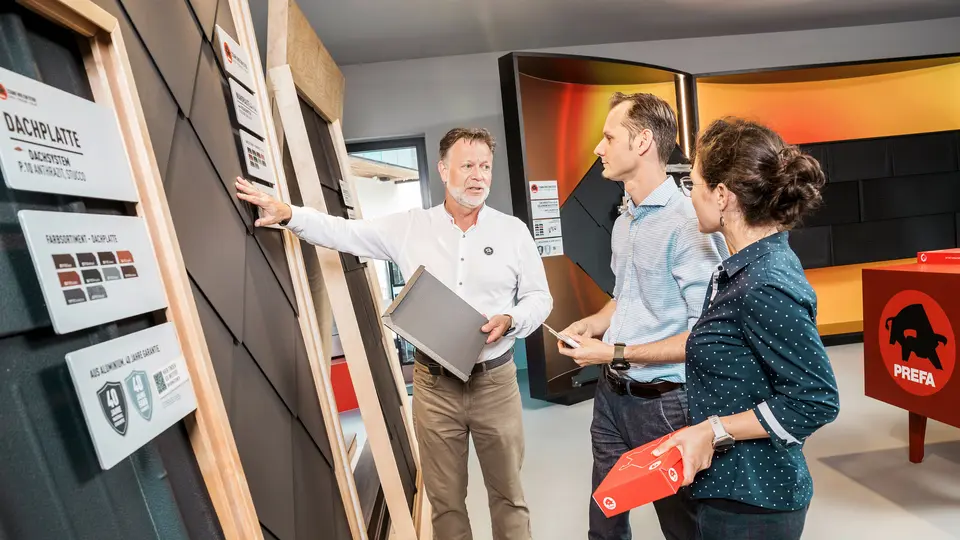 Ein Berater im PREFA Showroom zeigt zwei Kunden die verschiedenen Dachplattenmodelle. Er erklärt die Eigenschaften der Dachplatten in P.10 Anthrazit, während die Kunden aufmerksam zuhören und Prospektmaterialien in der Hand halten.