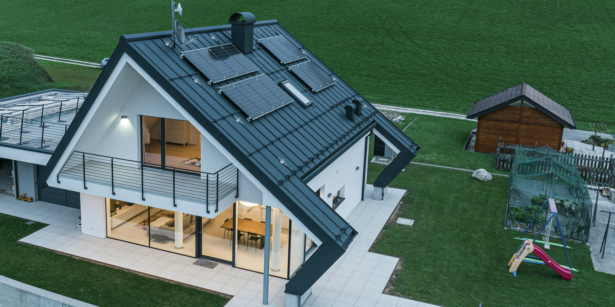 Modernes Einfamilienhaus in Borgo Valbelluna aus der Vogelperspektive in der Abenddämmerung. Im Haus erhellen Lichter die Wohnräume. Das markante Dach mit seinen außergewöhnlichen Auskragungen wurde mit einer robusten PREFALZ Stehfalzdeckung in P.10 Anthrazit eingedeckt. Das langlebige Aluminiumdach und die Photovoltaikmodule verbinden Nachhaltigkeit mit hochwertigem Design. Der markante Dachüberstand schützt Fassade und Terrasse vor Witterungseinflüssen. Große Glasflächen schaffen eine lichtdurchflutete Wohnatmosphäre, der umlaufende Balkon und die großzügige Terrasse maximieren den Wohnkomfort. PREFA Aluminium steht für Stabilität, Eleganz und Wartungsfreiheit.