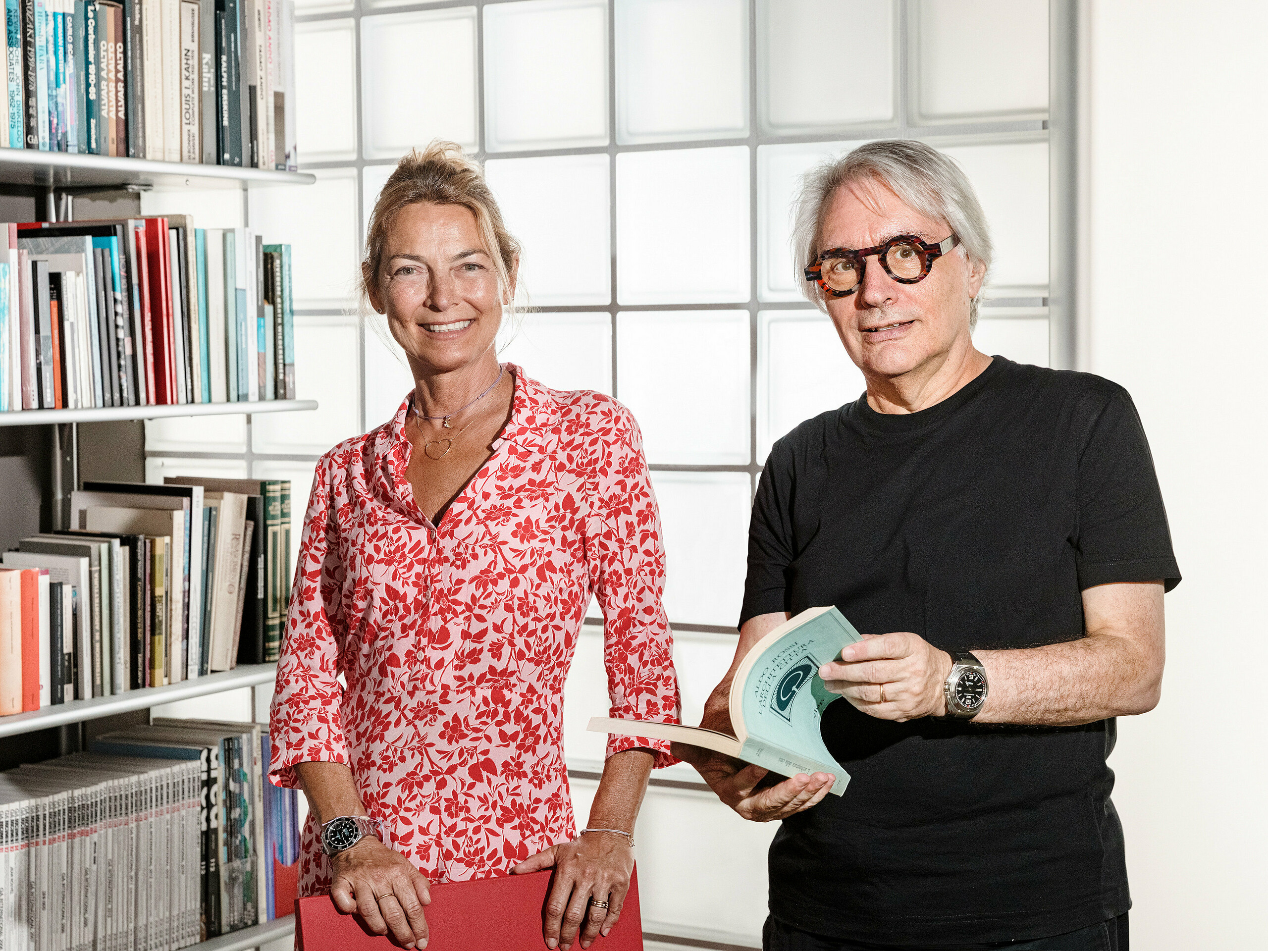 Der Bozener Architekt Claudio Lucchin und seine Büropartnerin Daniela Varnier posieren vor einer modernen Glasbausteinwand, die das Licht spannend bricht. Claudio Lucchin ist ganz in Schwarz gekleidet und hält ein Buch in den Händen. Daniela Varnier trägt ein knallrotes Top und lächelt freundlich. Dieses stilvolle Setting symbolisiert Innovation und Transparenz. Die Szene steht für zukunftsweisende Architektur, wie sie bei der Kaserne „Tempesti” in Corvara in Südtirol realisiert wurde.
