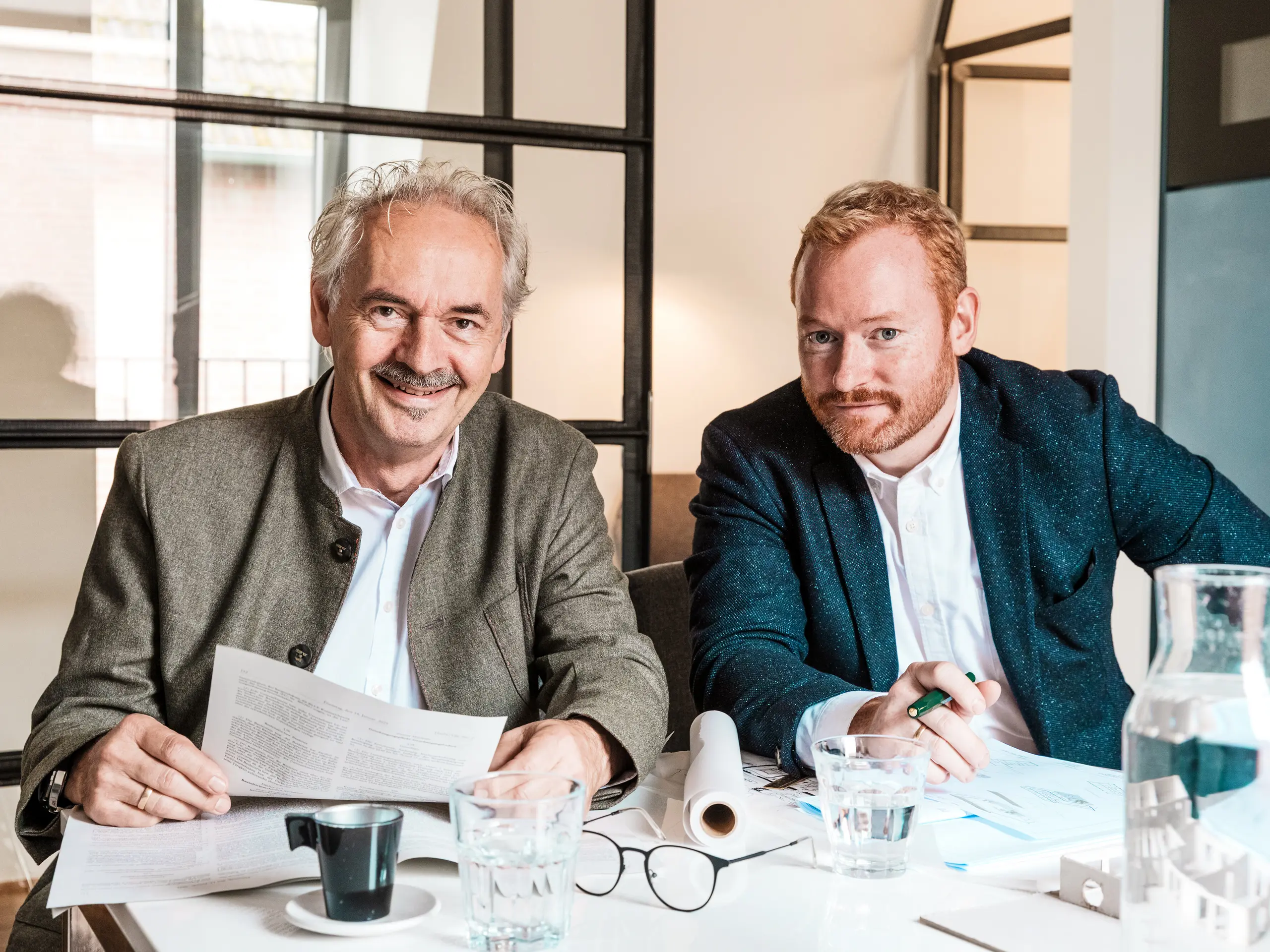 An einem Tisch sitzen zwei Architekten, die gerade Pläne erstellen. Vor ihnen stehen Getränke und ein Modell. Das Polizeikommissariat 43 in Hamburg, das von den beiden entworfen wurde, wurde mit PREFA Strangpressprofilen eindrucksvoll erweitert. Im Fokus der Szene stehen die beiden Herren von Pfluegelbauer Scheffczyk Architekten. Das Bild zeigt die Arbeit mit Plänen, Modellen und Unterlagen und spiegelt den langwierigen Weg zur Fertigstellung des Bauwerks wider.