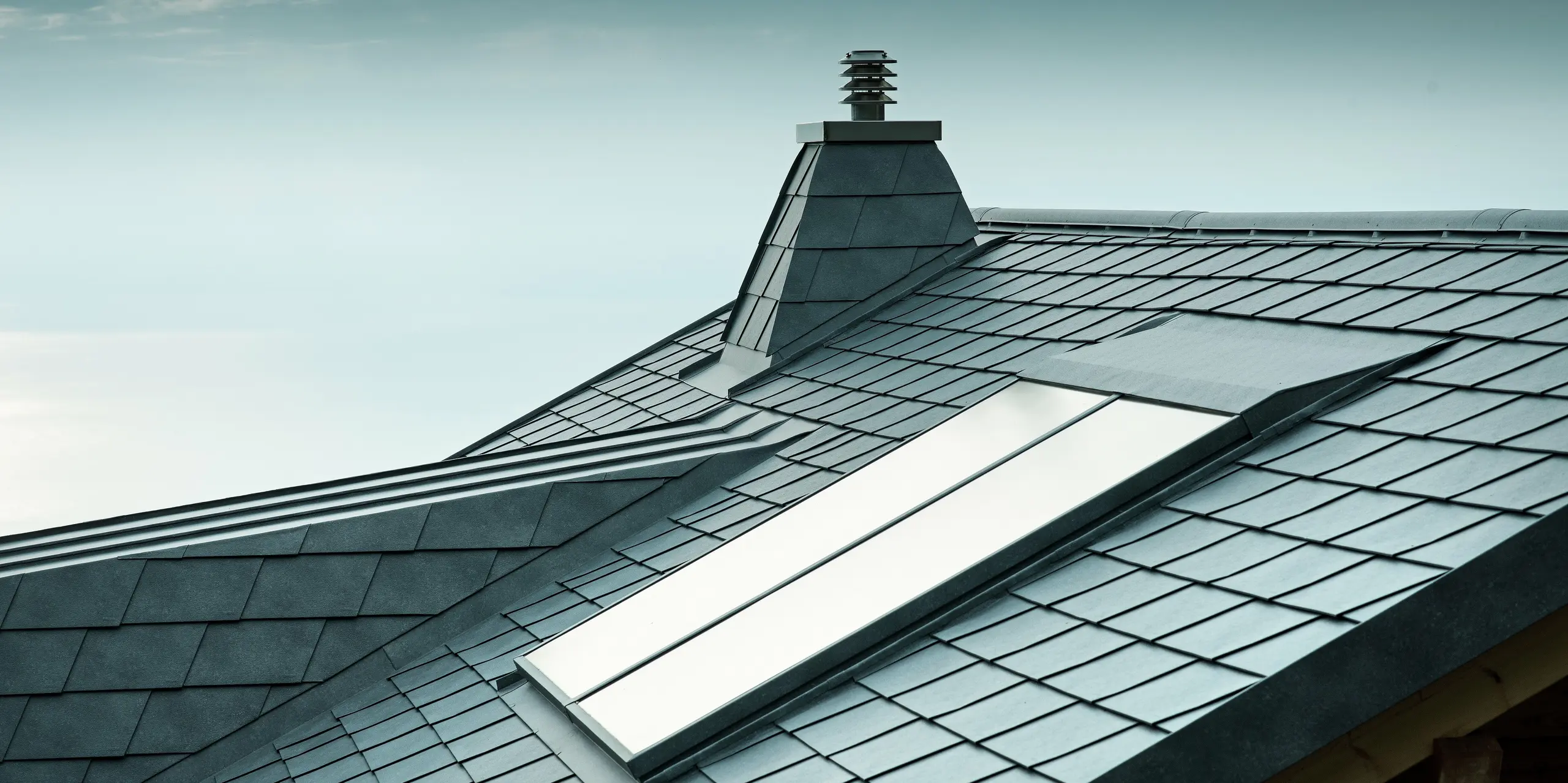Diese vielseitige Dachansicht zeigt steingraue PREFA-Dachschindeln sowie einen Kamin in Trapezform aus Aluminium. Das langlebige Aluminiumdach ist nicht nur stilvoll, sondern auch äußerst widerstandsfähig. Die exakte Ausführung der Dachgaube und des Dachfensters zeugt von der Detailgenauigkeit der PREFA-Verlegepartner.