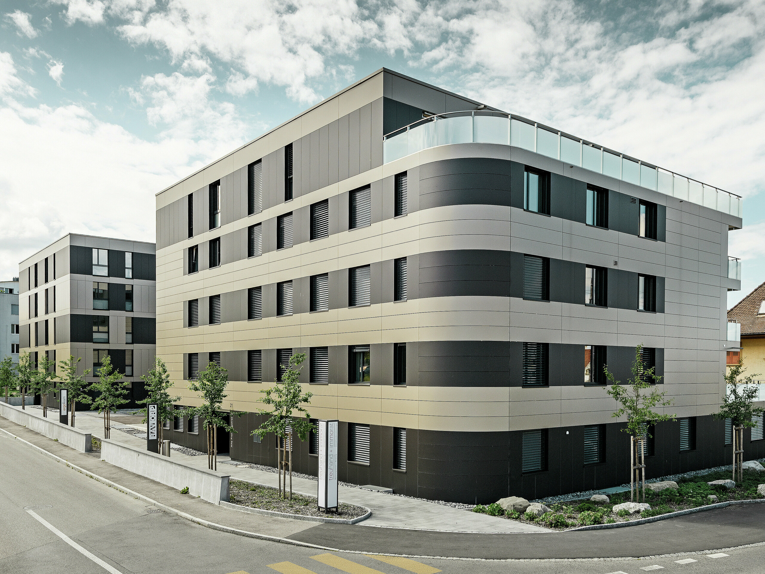 In der kubischen Wohnanlage von Murten (CH) sorgen die PREFA Aluminium Verbundplatten in Bronze und Schwarzgrau für eine optische Aufwertung und gleichzeitig für eine robuste und langlebige Fassade. Die Kombination dieser Farbtöne bietet ästhetische Akzente und schützt das Gebäude langfristig vor Wettereinflüssen. An der Gebäudeecke zur Straße hin ist eine Rundung zu erkennen, was das Rollbiegen der PREFABOND Aluminium Verbundplatten demonstrieren soll.