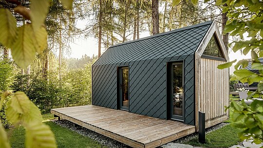 Ein minimalistisches Tiny House im Secret Koi Garden am Natterer See präsentiert sich eingebettet in eine ruhige Waldlandschaft. Dach und Fassade sind mit kleinteiligen PREFA Dach‑ und Wandrauten aus Aluminium in P.10 Anthrazit gestaltet, deren präzise, fischhautartige Struktur das Sonnenlicht sanft reflektiert. Die großzügige Holzterrasse öffnet den Raum zur Natur, während das langlebige, witterungsbeständige PREFA Aluminium die Architektur schützt und einen markanten Kontrast zu Holz und Grün schafft.