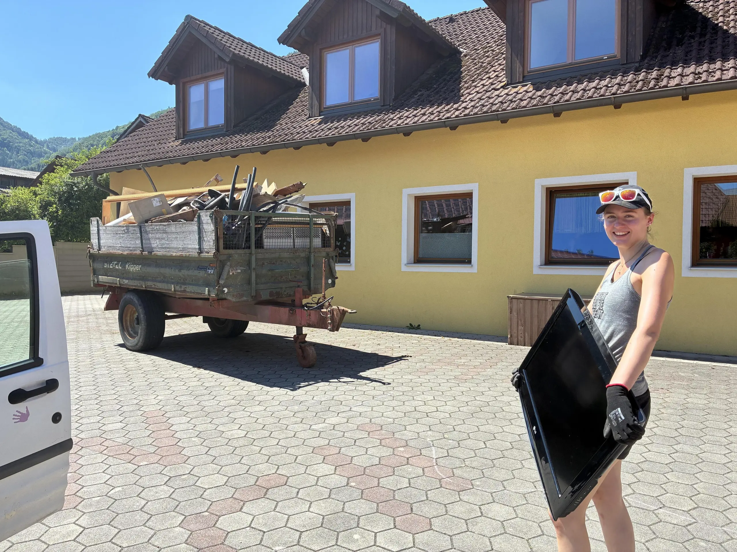 Tatkräftige Szene beim Social Team Tag am Kinderbauernhof Eschenau: Eine PREFA Mitarbeiterin trägt im Vordergrund einen alten Fernseher, während im Hintergrund ein Anhänger randvoll mit Sperrmüll und Holzresten beladen ist. Ein markanter Eyecatcher ist der strahlend blaue Himmel hinter dem ländlichen Gebäude. Das Bild vermittelt eindrucksvoll, wie PREFA Mitarbeitende durch gemeinsames Engagement konkrete Verbesserungen schaffen und soziale Einrichtungen mit vollem Einsatz nachhaltig unterstützen.