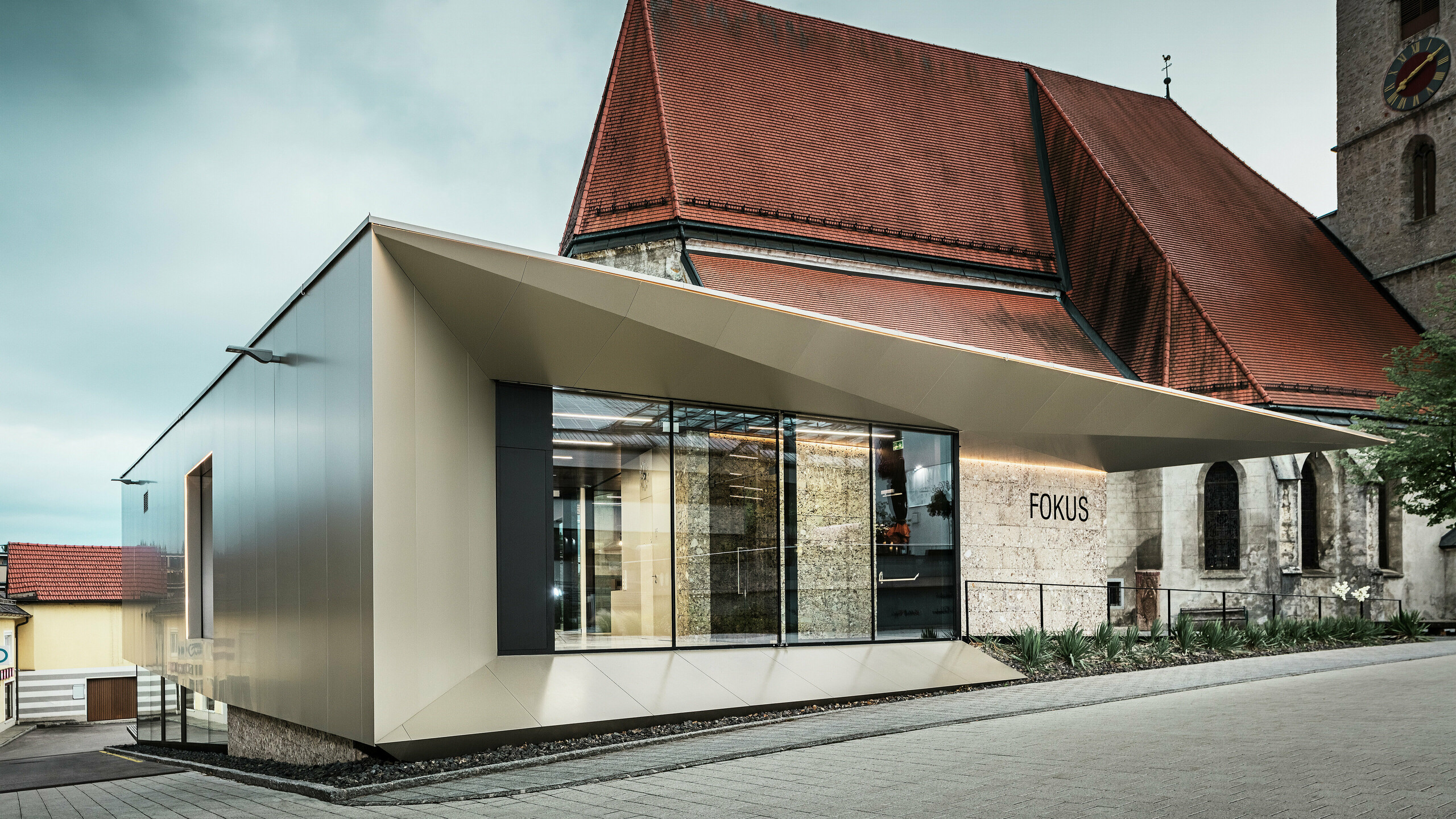 Im Vordergrund des Pfarrheims in Sierning glänzt die Aluminiumfassade durch ihre saubere Linienführung und hohe Formstabilität. Die Kombination aus Glasflächen und bronzefarbenen PREFA Aluminium Verbundplatten betont Offenheit, Beständigkeit und gestalterische Präzision bei gleichzeitiger Integration ins historische Umfeld.