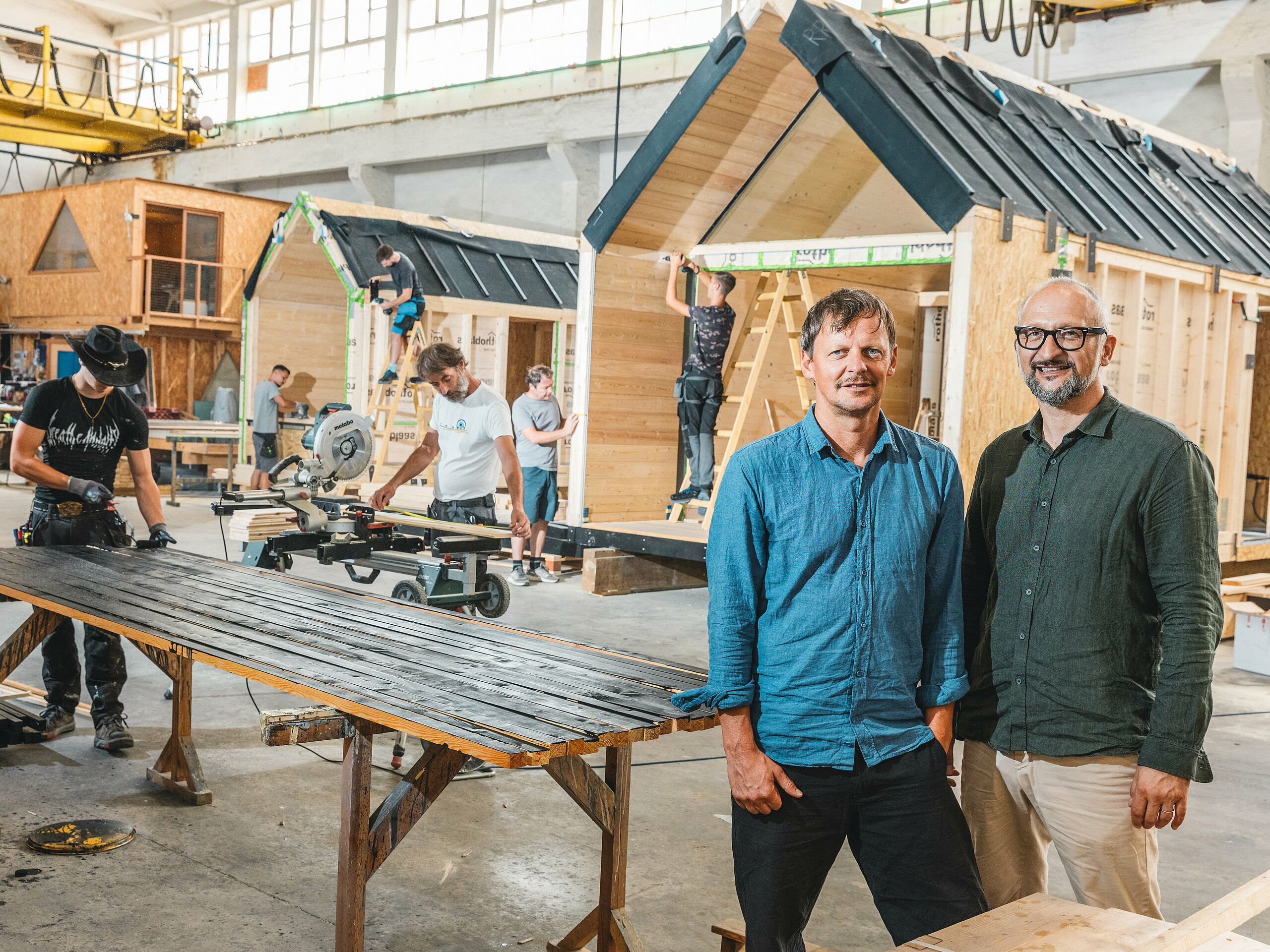 In einer Produktionshalle entstehen die minimalistischen Tiny Houses von Lushna. Das Unternehmen ist international erfolgreich und spezialisiert auf Minitekturen in Naturresorts. Im Vordergrund stehen die beiden Firmengründer: Architekt Peter Ličen und Marketingspezialist Jaka Ažman. Im Hintergrund werden A-Frame-Häuser montiert. Der markante Kontrast aus Holz und dunklem Aluminium macht die Architektur zum visuellen Eyecatcher, wovon der Secret Koi Garden am Natterer See zeugt.