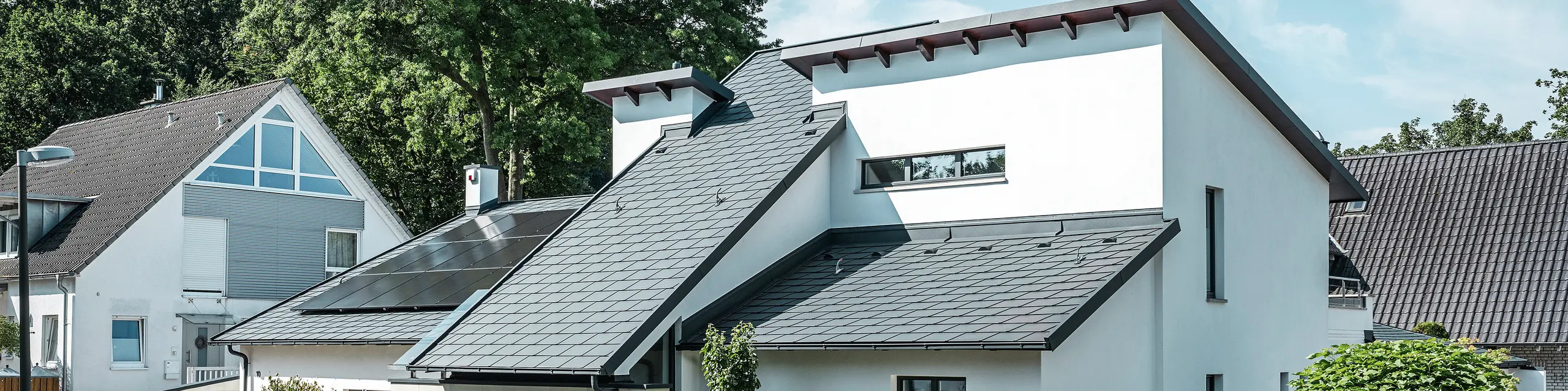 Modernes Einfamilienhaus mit weißer Putzfassade und dunklem PREFA Schrägdach aus Aluminium. Auf dem Dach sind integrierte Photovoltaikmodule und eine Aluminium-Dachentwässerung in Anthrazit zu sehen. Das Gebäude liegt in einer gepflegten Wohnsiedlung mit bepflanzten Vorgärten, gepflasterter Zufahrt und umliegenden Einfamilienhäusern.