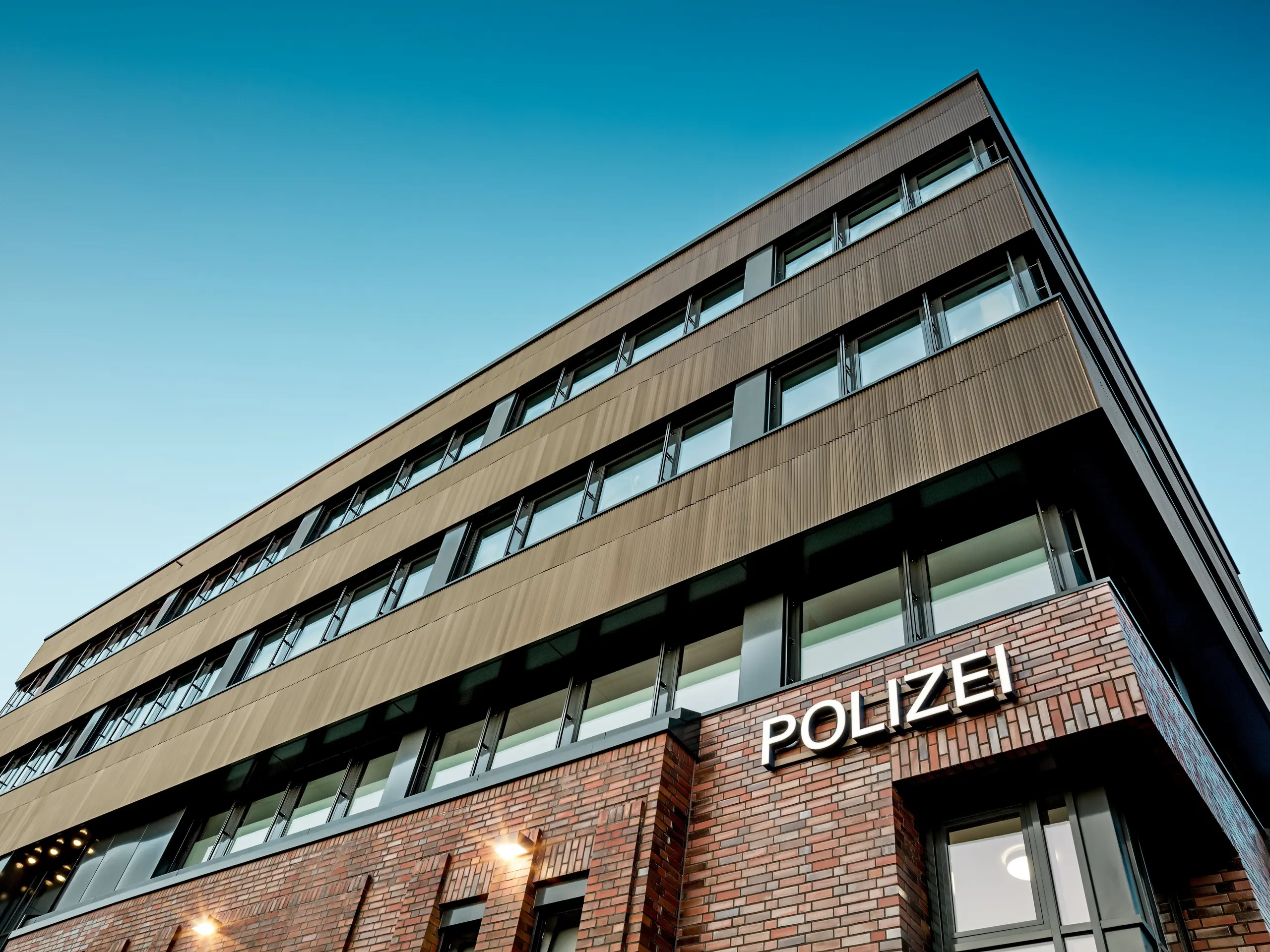 Architektonische Untersicht des Polizeikommissariats 43 in Hamburg: Über dem Backsteinbestand erhebt sich der präzise gegliederte Neubau mit einer PREFA Aluminiumfassade aus Strangpressprofilen „Zacke“ in edlem Mittelbronze. Die fein geriffelte Oberfläche wird zum visuellen Eyecatcher und erzeugt faszinierende Lichtreflexe entlang der klaren, horizontalen Fensterbänder. PREFA Aluminium steht dabei für höchste Korrosionsbeständigkeit, Langlebigkeit und minimale Wartung – eine nachhaltige, designstarke Lösung für moderne und funktionale Fassadenarchitektur.