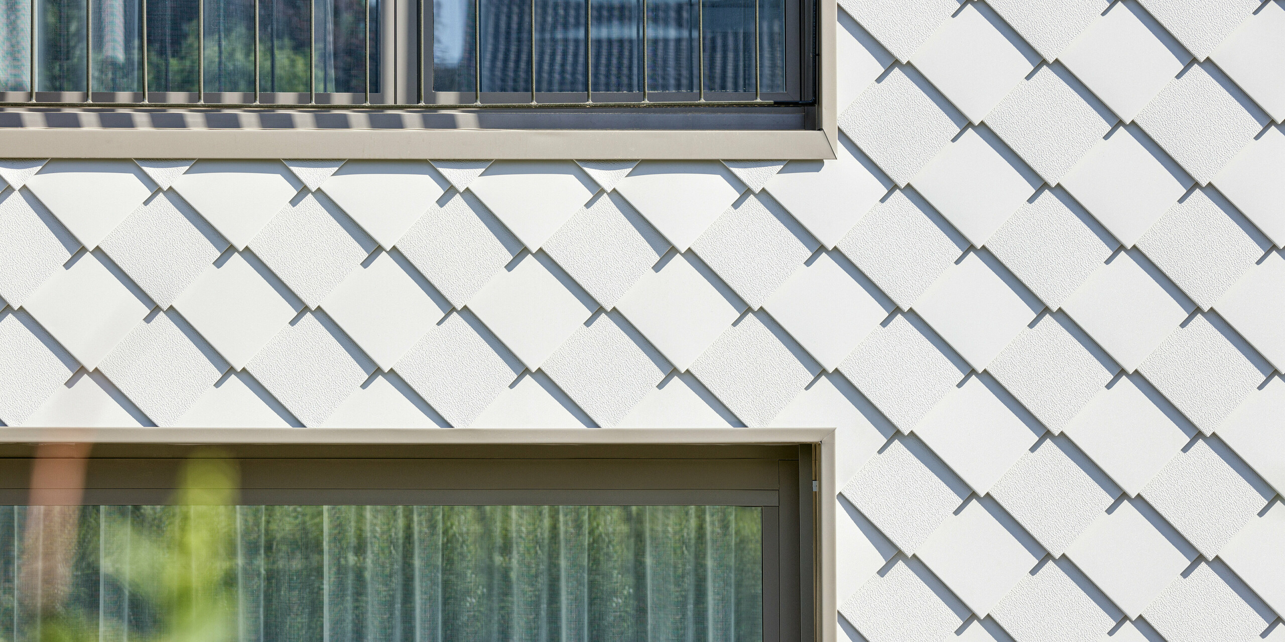 Detailansicht der modernen Fassadengestaltung eines Mehrfamilienhauses in Wiesendangen, CH. Die Gebäudehülle besteht aus PREFA Wandrauten 20x20 in P.10 Prefaweiß, die strukturierte Eleganz und zeitgenössisches Design vermitteln. Das Bild zeigt die präzise Montage der Aluminiumrauten, die zwischen zwei Fenstern mit französischem Balkon liegen.