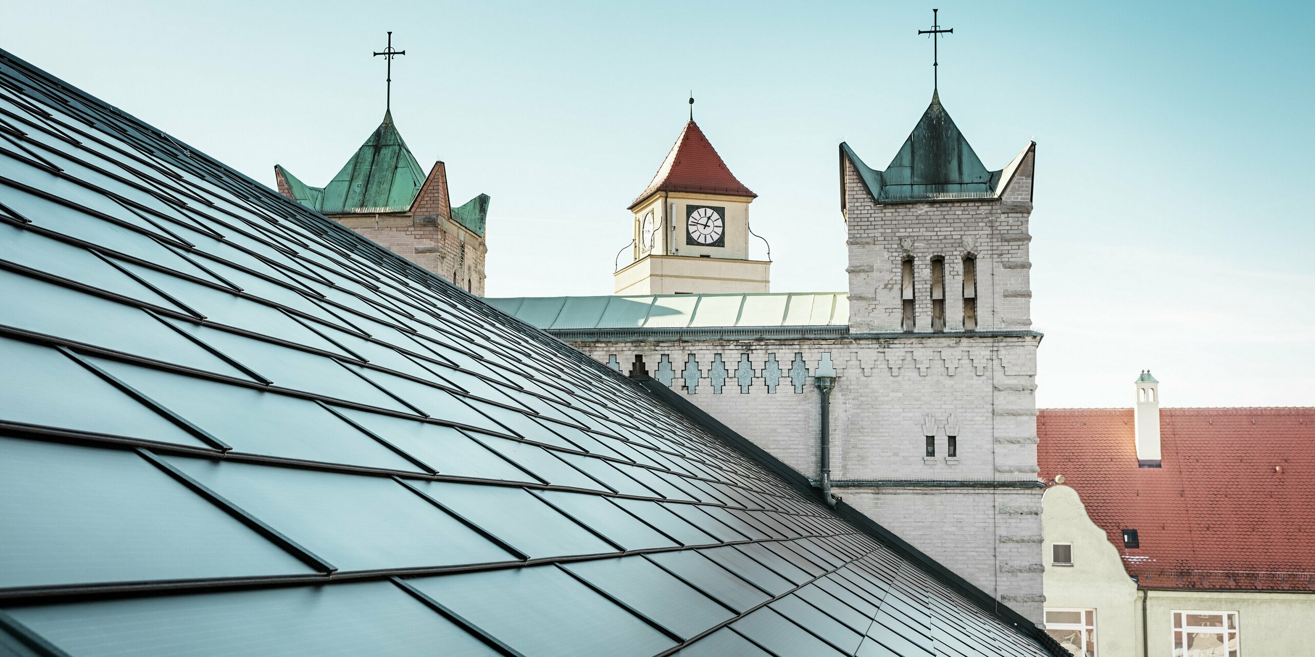 Das prägnante Dach der St. Karl Borromäus Kirche in Nürnberg wurde im Zuge einer umfassenden Sanierung mit schwarzen PREFA-Solardachplatten bedeckt. Der Detailblick auf das Satteldach und die markanten Kirchtürme im Hintergrund vermittelt die gelungene Einbindung moderner Solartechnologie in die historische Architektur der Kirche. Die Solardachplatten sorgen für nachhaltige Energie und fügen sich nahtlos in das traditionelle Erscheinungsbild ein. Dies ist ein weiteres Beispiel für die gelungene Verbindung von Denkmalschutz und innovativer Energiegewinnung. Im Hintergrund ist ein strahlend blauer Himmel zu erkennen.