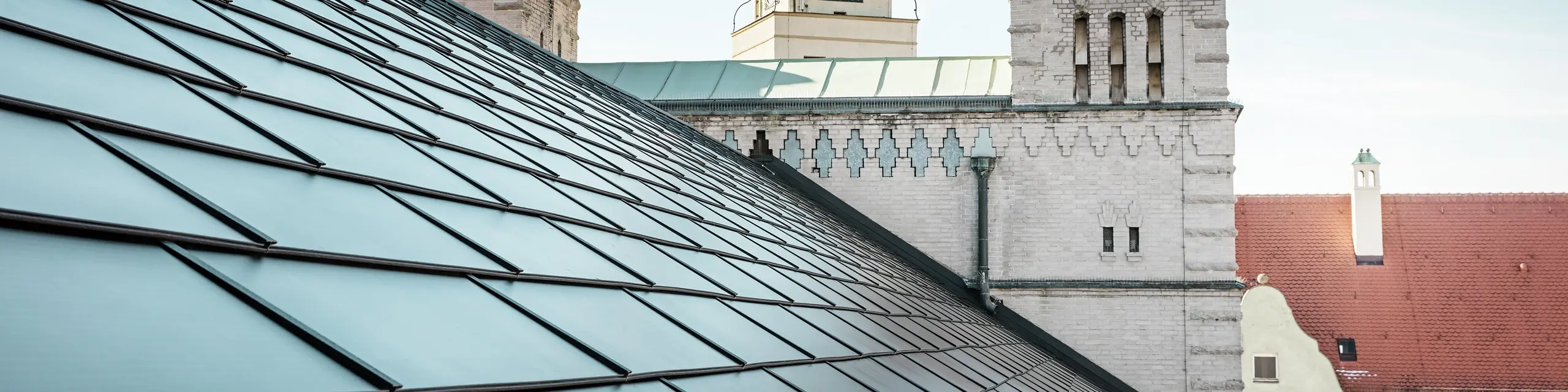 Das prägnante Dach der St. Karl Borromäus Kirche in Nürnberg wurde im Zuge einer umfassenden Sanierung mit schwarzen PREFA-Solardachplatten bedeckt. Der Detailblick auf das Satteldach und die markanten Kirchtürme im Hintergrund vermittelt die gelungene Einbindung moderner Solartechnologie in die historische Architektur der Kirche. Die Solardachplatten sorgen für nachhaltige Energie und fügen sich nahtlos in das traditionelle Erscheinungsbild ein. Dies ist ein weiteres Beispiel für die gelungene Verbindung von Denkmalschutz und innovativer Energiegewinnung. Im Hintergrund ist ein strahlend blauer Himmel zu erkennen.