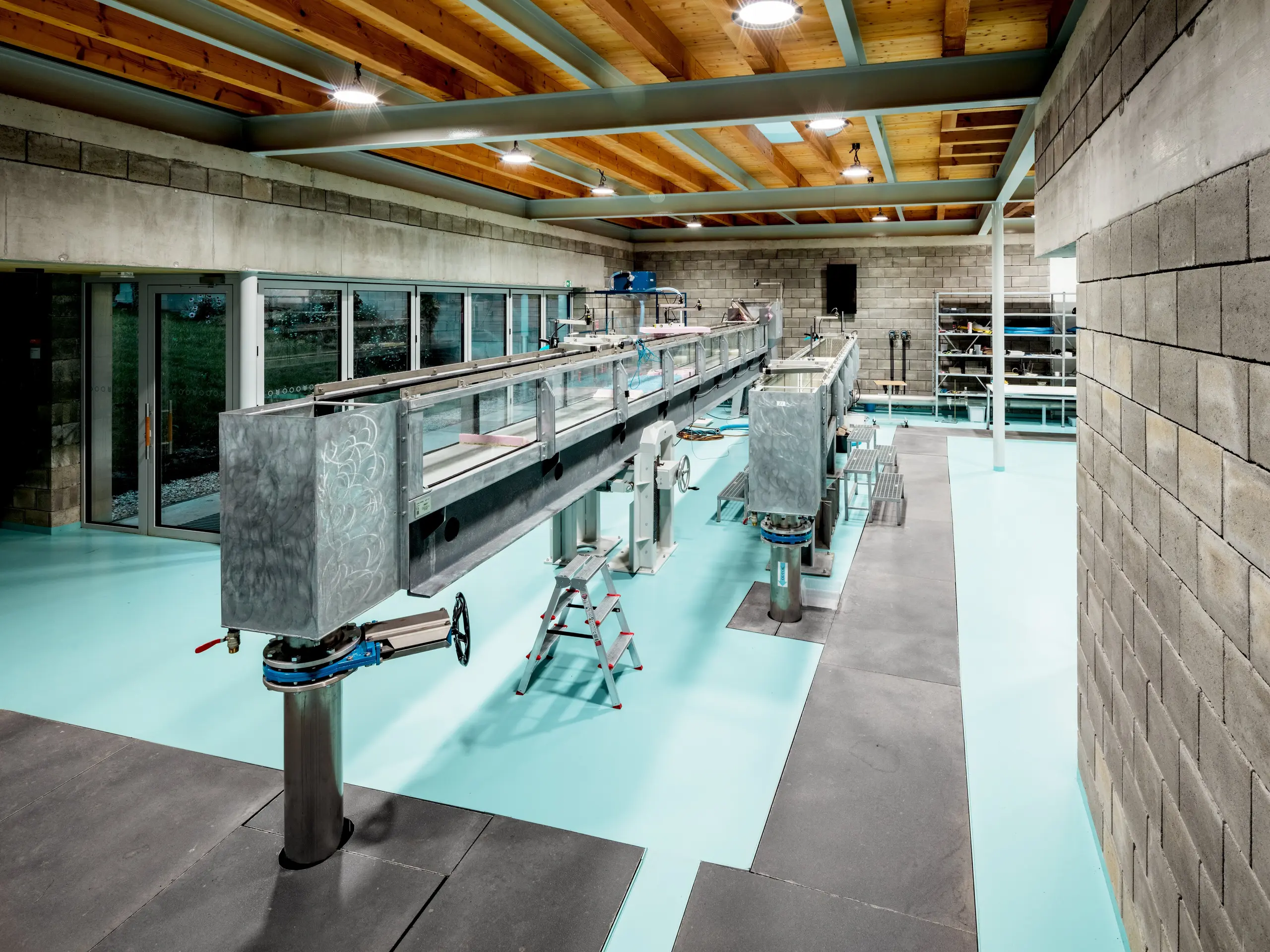 Das moderne Hydrauliklabor im Envirocenter Nitra beeindruckt mit zwei langen Strömungskanälen für wissenschaftliche Experimente rund um Wasserbewegungen. Sichtbare Stahlträger, eine warme Holzdecke, rohe Betonsteinwände und der türkisfarbene Boden prägen die offene Forschungslandschaft. Großzügige Glasfronten schaffen eine helle Atmosphäre und verbinden innovative Umweltforschung mit zeitgemäßer Architektur und bestehender Bausubstanz.