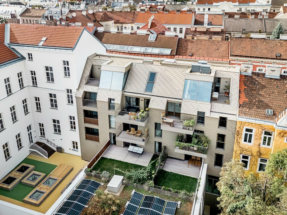 Die Vogelperspektive zeigt eine beeindruckende Aussicht auf die Erweiterung eines Wohngebäudes in Wien, bei dem PREFA Rauten 29×29 in Bronze auf der Dach- und Fassadengestaltung verwendet wurden. Die modernen Rauten harmonieren perfekt mit der traditionellen Architektur der Umgebung und schaffen eine attraktive visuelle Verbindung zwischen Alt und Neu. Im Innenhof ist ein durchdachtes Design mit Solarzellen, grünen Flächen und Holzterrassen zu sehen, das den nachhaltigen Ansatz der Wohnanlage unterstreicht. Die lebendige Mischung aus urbanem Raum und Natur wird durch die Dachterrassen und Balkone ergänzt, die den Bewohnern einen exklusiven Blick über die Wiener Dächer bieten.