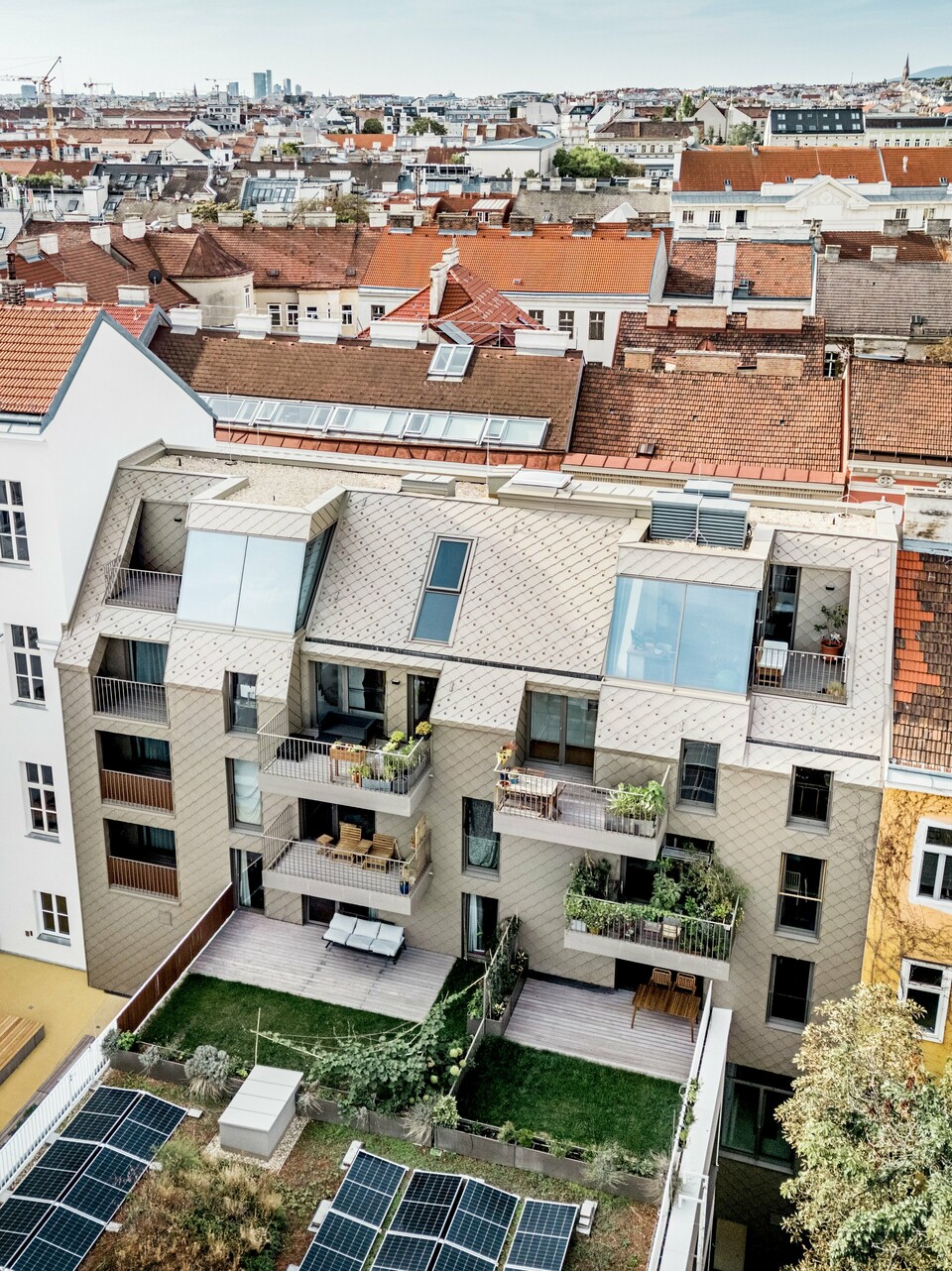 Die Vogelperspektive zeigt eine beeindruckende Aussicht auf die Erweiterung eines Wohngebäudes in Wien, bei dem PREFA Rauten 29×29 in Bronze auf der Dach- und Fassadengestaltung verwendet wurden. Die modernen Rauten harmonieren perfekt mit der traditionellen Architektur der Umgebung und schaffen eine attraktive visuelle Verbindung zwischen Alt und Neu. Im Innenhof ist ein durchdachtes Design mit Solarzellen, grünen Flächen und Holzterrassen zu sehen, das den nachhaltigen Ansatz der Wohnanlage unterstreicht. Die lebendige Mischung aus urbanem Raum und Natur wird durch die Dachterrassen und Balkone ergänzt, die den Bewohnern einen exklusiven Blick über die Wiener Dächer bieten.