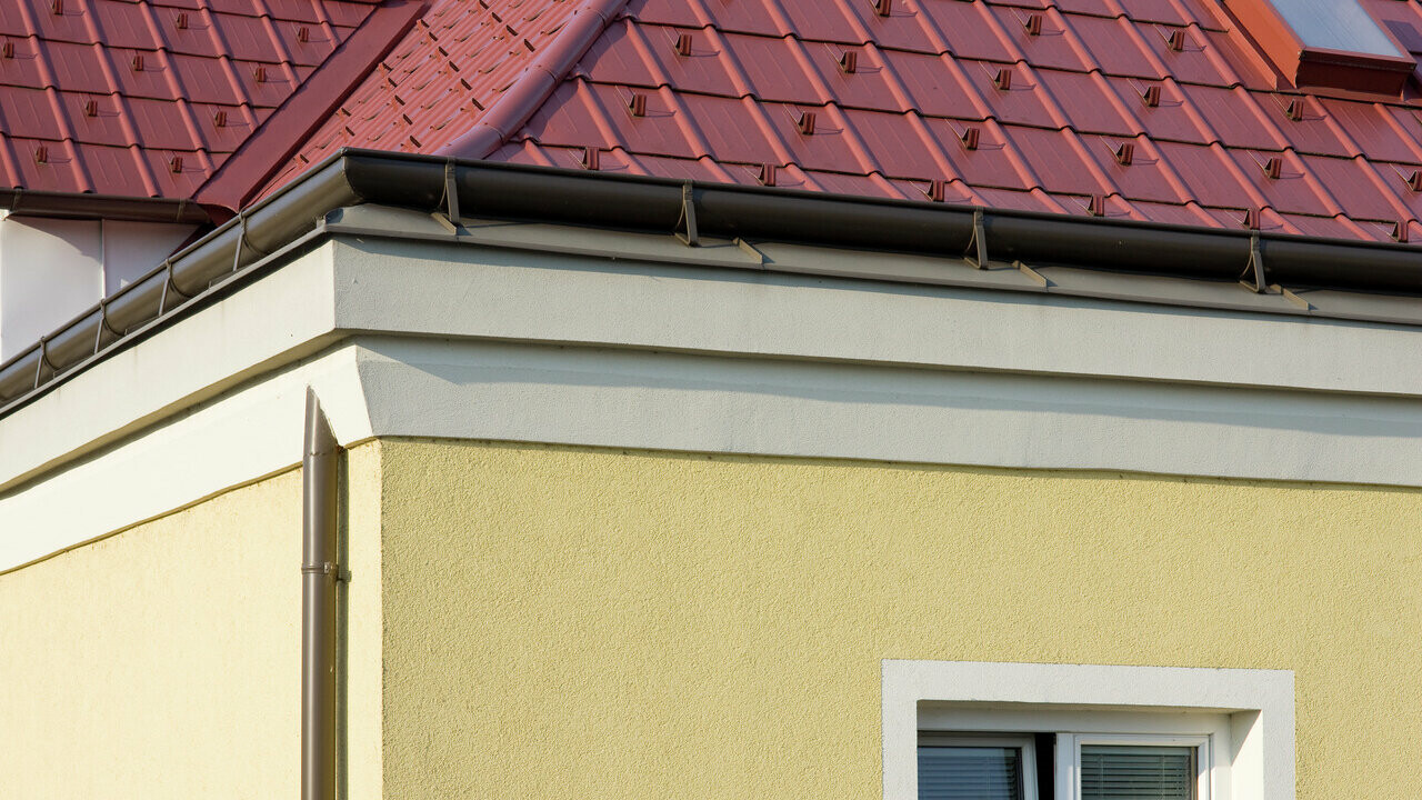 Hauseck mit gelber Putzfassade und brauner PREFA Saumrinne (Aufdachrinne) mit rotem PREFA Dachplattendach