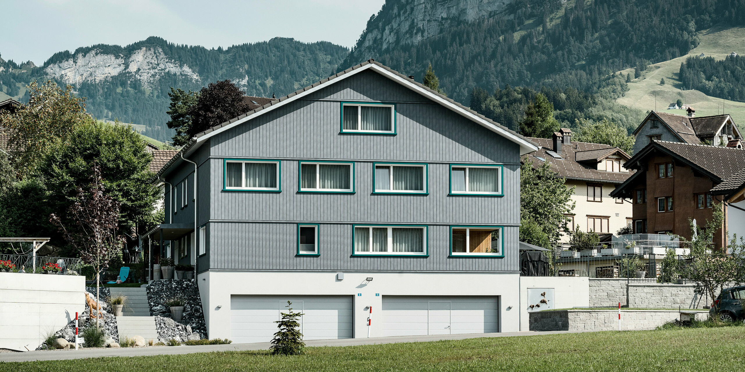 Vollansicht eines Mehrfamilienhauses in Weissbad, Schweiz, mit einer PREFA Siding Fassade in P.10 Steingrau. Die vertikal verlegten Aluminiumsidings verleihen dem Gebäude ein modernes, strukturiertes Aussehen, das sich von der malerischen Kulisse der Schweizer Alpen abhebt. Die türkisfarbenen Akzente der Fensterrahmen fügen eine lebendige Note hinzu, während die robuste Bauweise der PREFA Produkte die Beständigkeit gegenüber den alpinen Wetterbedingungen verspricht.