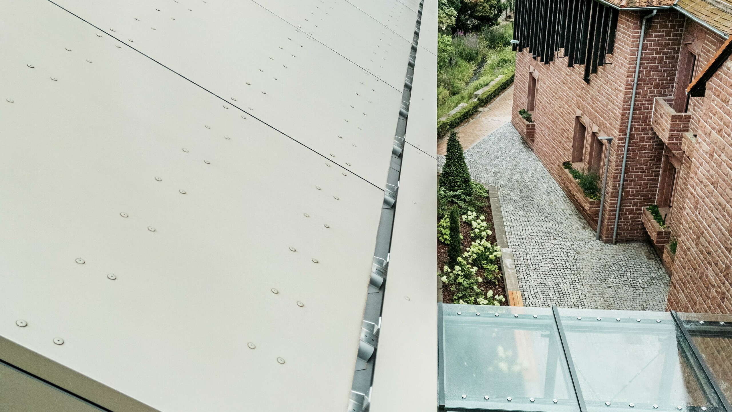 Detailaufnahme des Daches des Rathauses in Alzenau, das mit Aluminium-Verbundplatten von PREFA in einem sanften Bronze-Ton gedeckt ist. Die Platten sind präzise verlegt und mit sichtbaren Befestigungselementen versehen, was der Fassade einen modernen, industriellen Look verleiht. Im Vordergrund ist ein gläserner Verbindungsgang zu sehen, der das Rathaus mit einem benachbarten Gebäude verbindet.