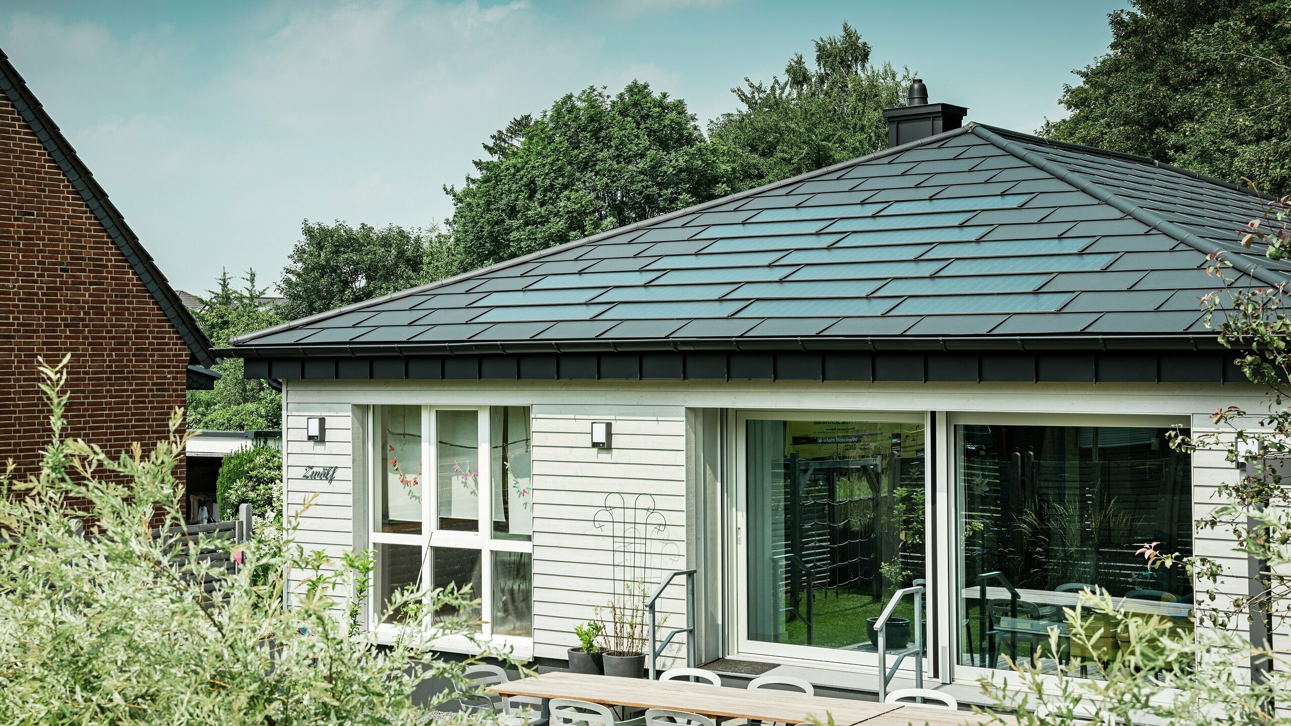 Sanierter Bungalow in Rommerskirchen mit elegantem PREFA Aluminiumdach. PREFA Solardachplatten und Dachplatten R.16 in P.10 Schwarz schützen dauerhaft und erzeugen nachhaltig Strom aus Sonnenenergie. Die großzügige Fensterfront sorgt für viel Licht, die Dachentwässerung mit Saumrinne und Ablaufrohr verbindet Funktionalität und Design perfekt.