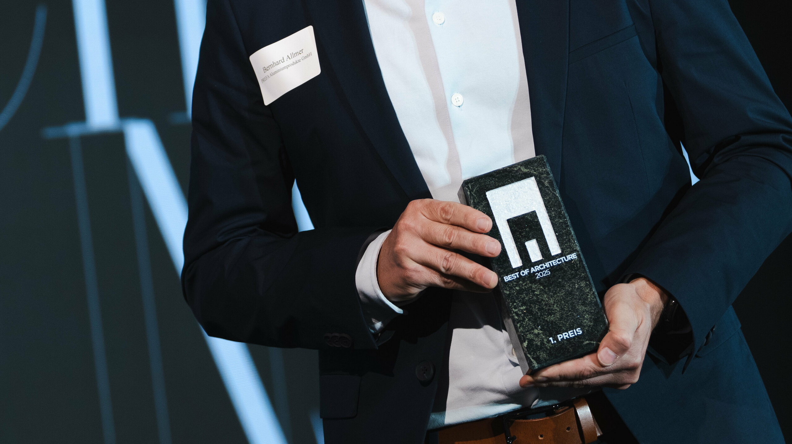 Preisverleihung beim renommierten Callwey-Wettbewerb ‚Wohnbauten des Jahres‘ 2025: Im Vordergrund eine elegante Person in dunklem Anzug und weißem Hemd, die stolz die Auszeichnung ‚Architects’ Choice‘ präsentiert. Die Person ist Bernhard Allmer, der Produktverantwortliche für die PREFA Solarprodukte. Der Award besteht aus einer edlen, dunklen Steinplatte mit modernem Logo und klarer Typografie. Im Hintergrund ist eine stilvolle Bühne mit großformatiger Schrift und akzentuierter Beleuchtung zu sehen, die den feierlichen Charakter des Events unterstreicht. Die Szene vermittelt Innovation und Exzellenz im Bereich Architektur.