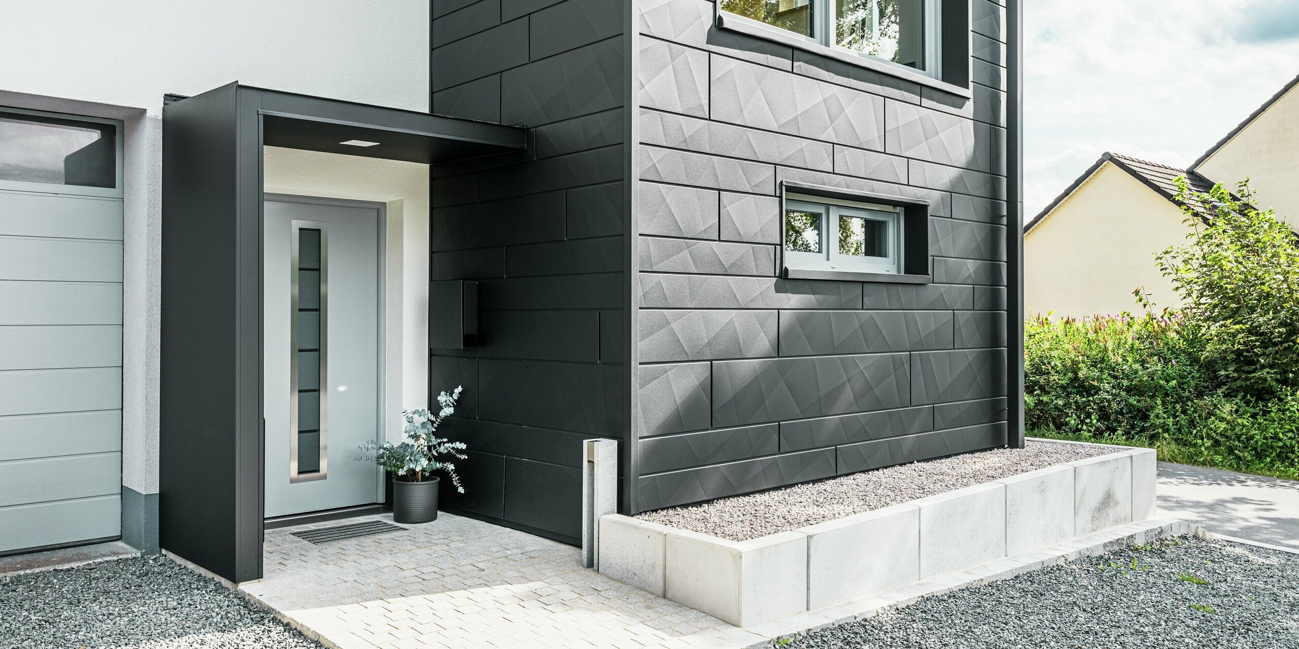 Die elegante PREFA Siding.X Fassade in P.10 Schwarz dominiert den Eingangsbereich des modernisierten Einfamilienhauses in Teuschnitz. Die hochwertige Blechfassade verleiht dem Gebäude einen modernen und zeitlosen Charakter. Die klar strukturierten Fassadenpaneele schaffen ein markantes, geometrisches Design, während die schwarzen Aluminium Verbundplatten die Eingangstür und die Fenster umrahmen. Das PREFA Quadratrohr fügt sich nahtlos in das anspruchsvolle Design ein und unterstreicht die funktionale und ästhetische Wirkung der Sanierung. Der Weg zum Haus ist mit Pflastersteinen und Kies versehen.