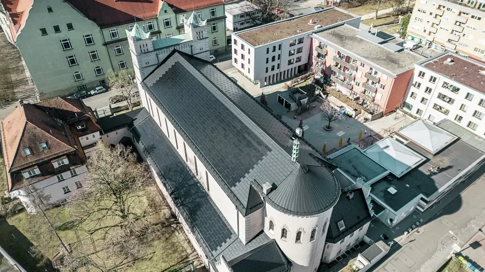 Vogelperspektive auf die Rückseite der St. Karl Borromäus Kirche in Nürnberg. Die schwarzen PREFA Solardachplatten decken das markante, runde Satteldach über dem Hauptteil der Kirche als auch das Dach des Nebengebäudes. Die Aluminiumdachplatten mit integrierten PV-Modulen fügen sich perfekt in das historische Erscheinungsbild der Kirche ein. Der Blick auf das moderne Umfeld mit angrenzenden Wohngebäuden und dem grünen Hofbereich verdeutlicht die gelungene Verbindung von Denkmalschutz, moderner Architektur und nachhaltiger Energiegewinnung. Eine ideale Lösung für den Denkmalschutz im urbanen Raum.