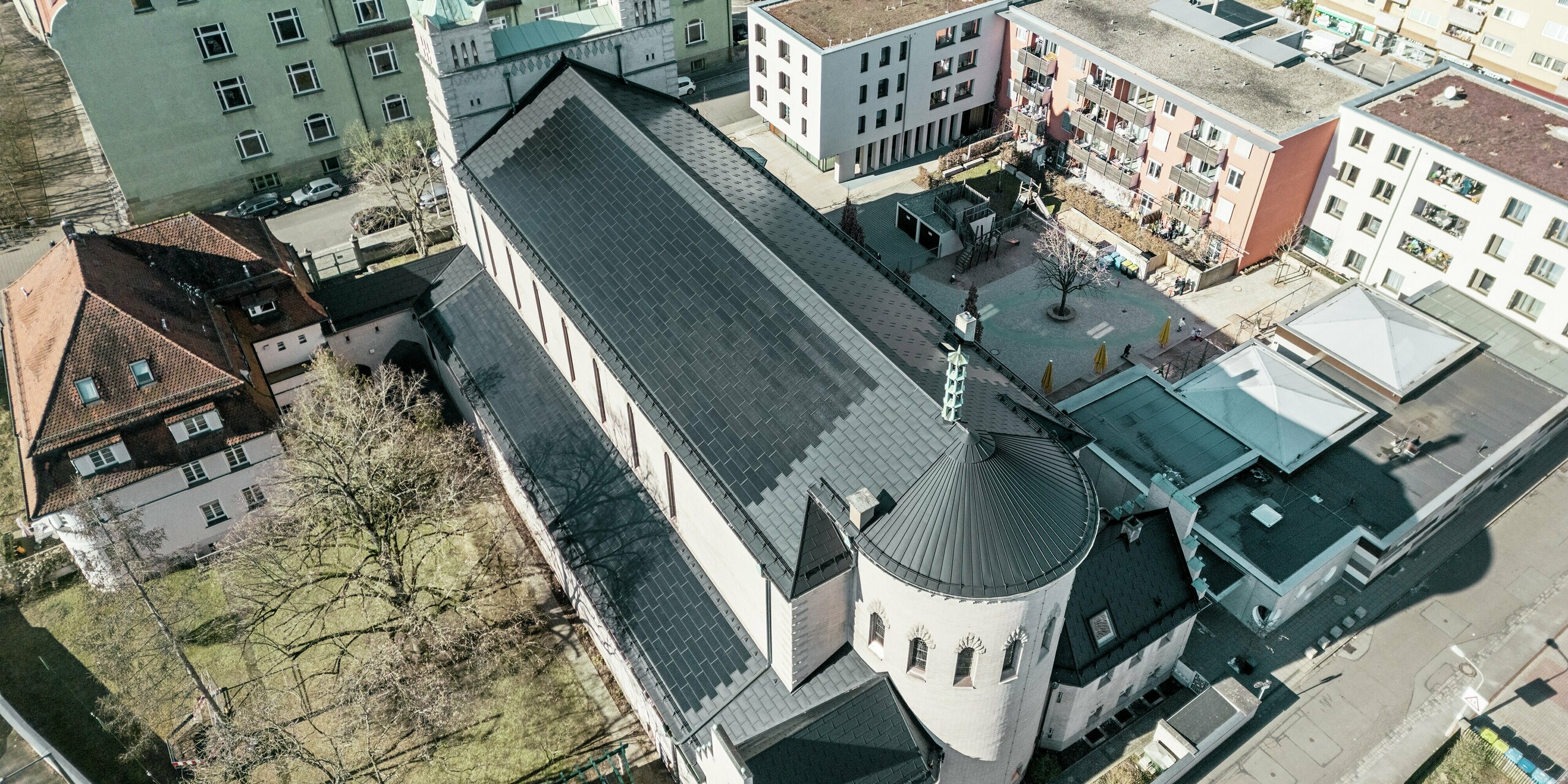 Vogelperspektive auf die Rückseite der St. Karl Borromäus Kirche in Nürnberg. Die schwarzen PREFA Solardachplatten decken das markante, runde Satteldach über dem Hauptteil der Kirche als auch das Dach des Nebengebäudes. Die Aluminiumdachplatten mit integrierten PV-Modulen fügen sich perfekt in das historische Erscheinungsbild der Kirche ein. Der Blick auf das moderne Umfeld mit angrenzenden Wohngebäuden und dem grünen Hofbereich verdeutlicht die gelungene Verbindung von Denkmalschutz, moderner Architektur und nachhaltiger Energiegewinnung. Eine ideale Lösung für den Denkmalschutz im urbanen Raum.
