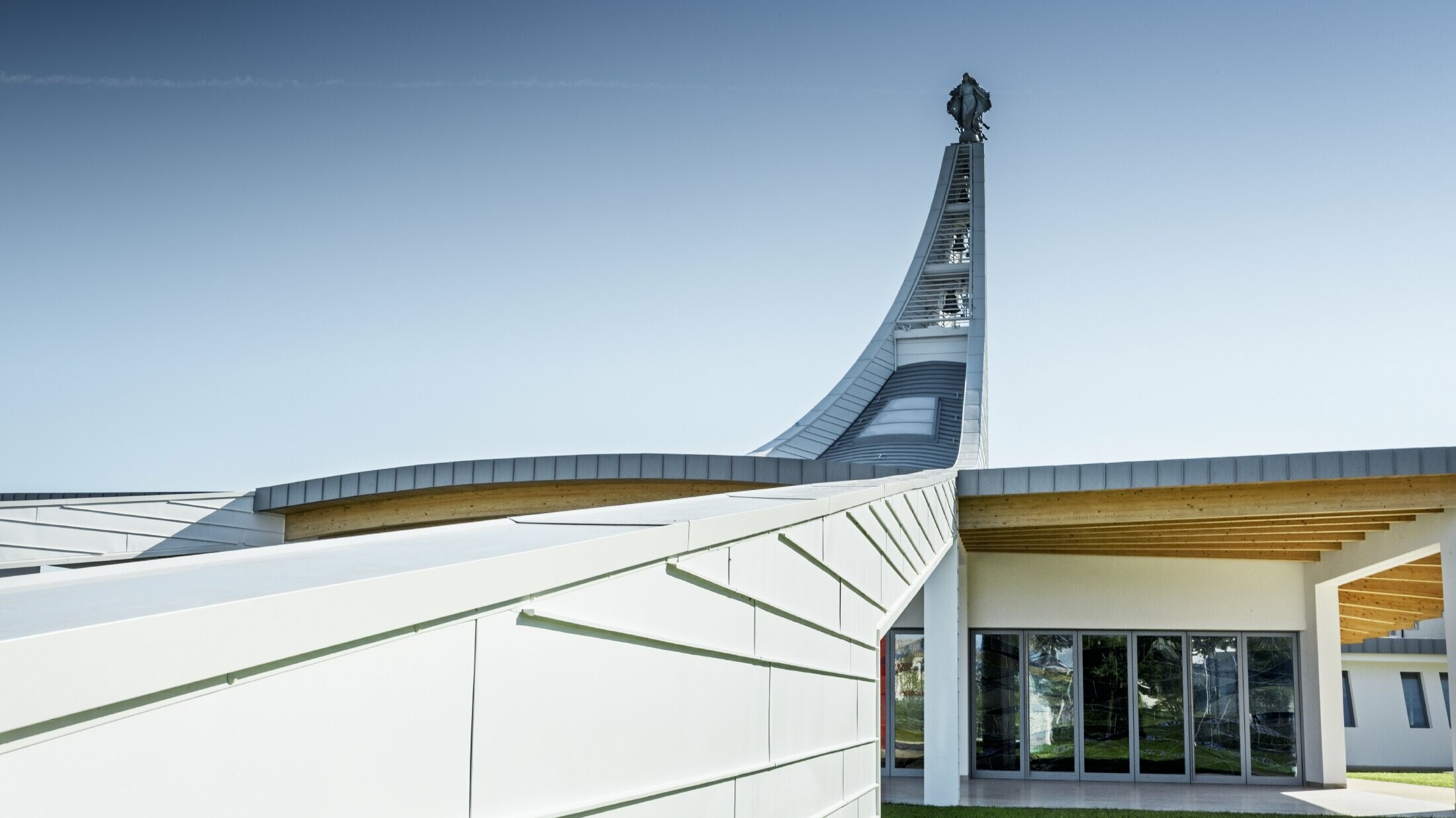 Modernes Kirchengebäude mit geschwungener Dachkonstruktion aus hellen Aluminiumpaneelen. Im Hintergrund erhebt sich ein hoher, elegant gebogener Turm mit einer Statue an der Spitze. Die Architektur kombiniert klare Linien mit offenen Glasflächen und Holzelementen unter dem Dach. Der Himmel ist wolkenlos und betont die helle, futuristische Gestaltung des Bauwerks