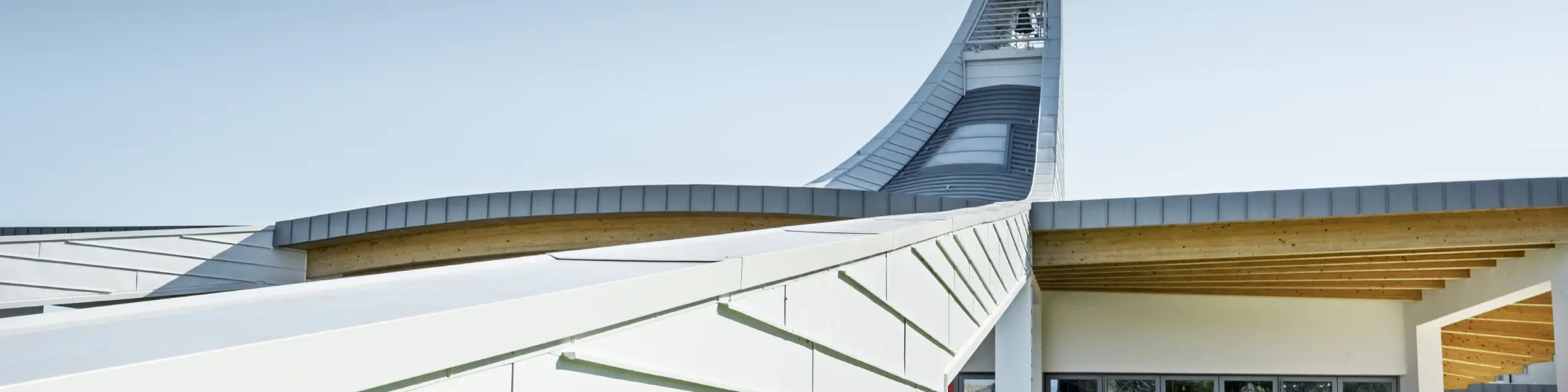 Modernes Kirchengebäude mit geschwungener Dachkonstruktion aus hellen Aluminiumpaneelen. Im Hintergrund erhebt sich ein hoher, elegant gebogener Turm mit einer Statue an der Spitze. Die Architektur kombiniert klare Linien mit offenen Glasflächen und Holzelementen unter dem Dach. Der Himmel ist wolkenlos und betont die helle, futuristische Gestaltung des Bauwerks
