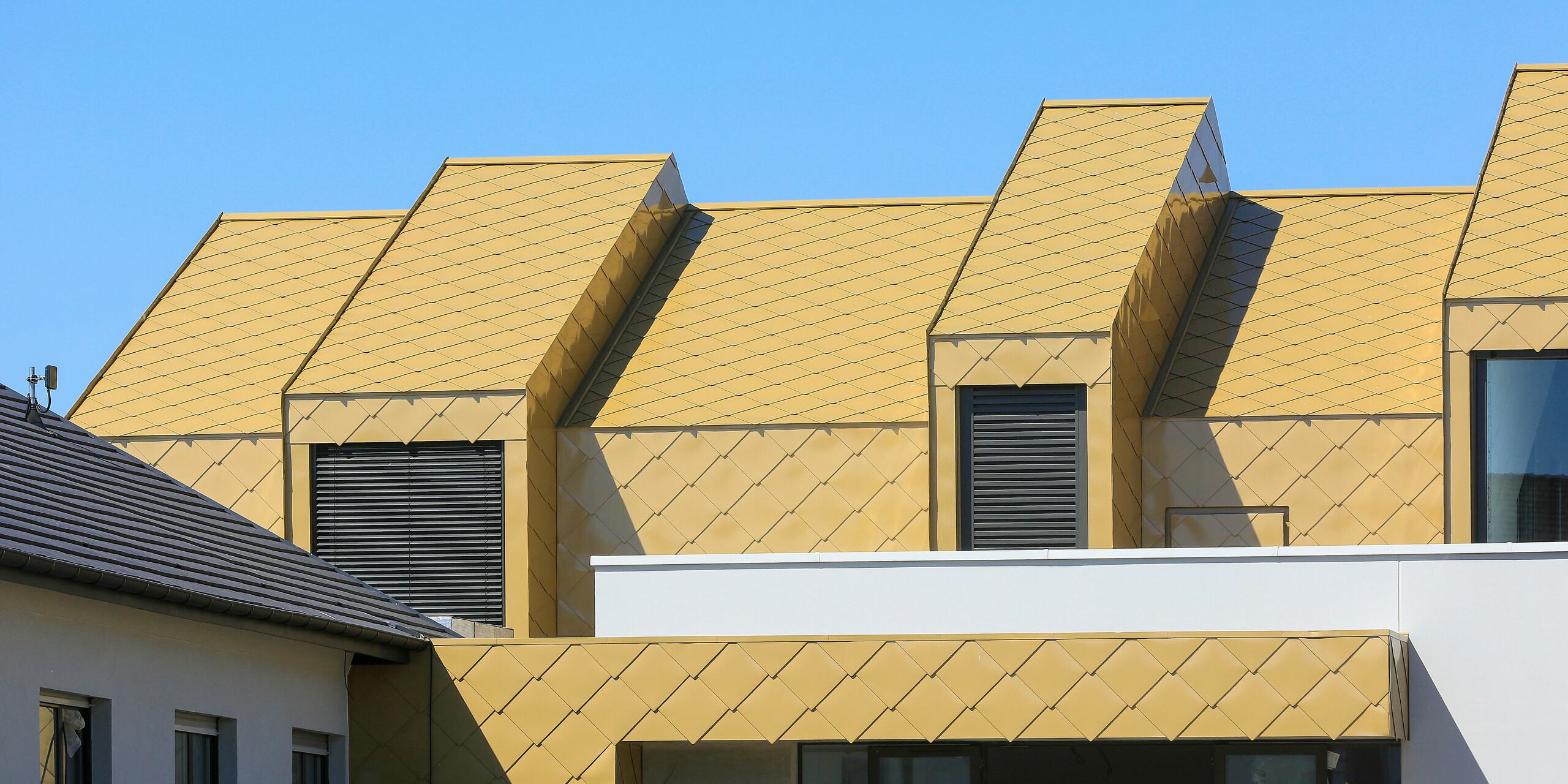 Nahaufnahme des Balkons und der Dachfenster am Gemeinde- und Kulturzentrum Fessenheim in Frankreich. Das Dach sowie Teile der Fassade wurden mit PREFA Dach- und Wandrauten 44 × 44 in Sparkling Gold verkleidet.