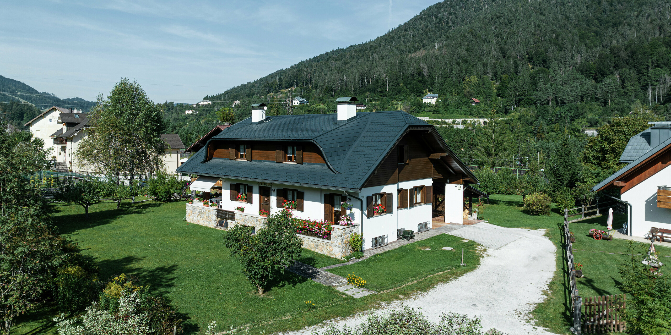 Dieses sanierte Einfamilienhaus in Tarvis (Italien) besticht durch sein harmonisches Design und das hochwertige PREFA Aluminiumdach. Die Hechtgaube verleiht dem Dach eine elegante Form und die widerstandsfähigen PREFA Dachschindeln in P.10 Anthrazit schützen das Gebäude zuverlässig vor Witterungseinflüssen. Die Kombination aus weißen Fassaden, dunklen Holzelementen und der gepflegten Gartenanlage unterstreicht den traditionellen, alpinen Charakter des Hauses inmitten einer malerischen Berglandschaft.