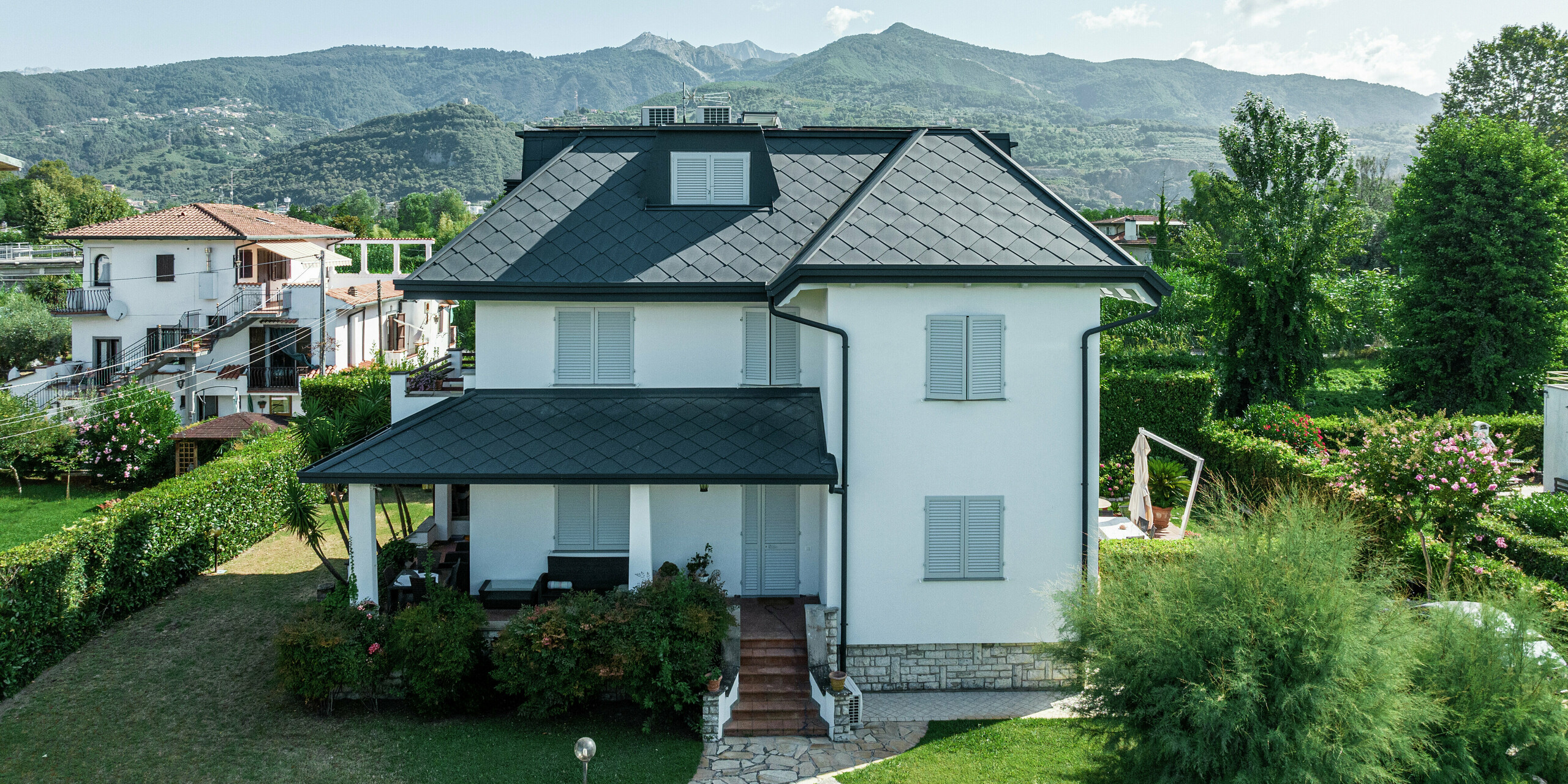 Frontansicht eines schlichten Einfamilienhauses in Montignoso, Italien. Das Dach mit der PREFA Dachraute 44 × 44 in P.10 Steingrau kommt vor dem strahlend blauen Himmel besonders gut zur Geltung. Die hochwertige Aluminiumdeckung sorgt für eine moderne, elegante Optik und bietet gleichzeitig höchsten Schutz vor extremen Witterungseinflüssen. Die Kombination aus weißer Fassade, dunklem Dach und der grünen Umgebung schafft ein harmonisches Gesamtbild.