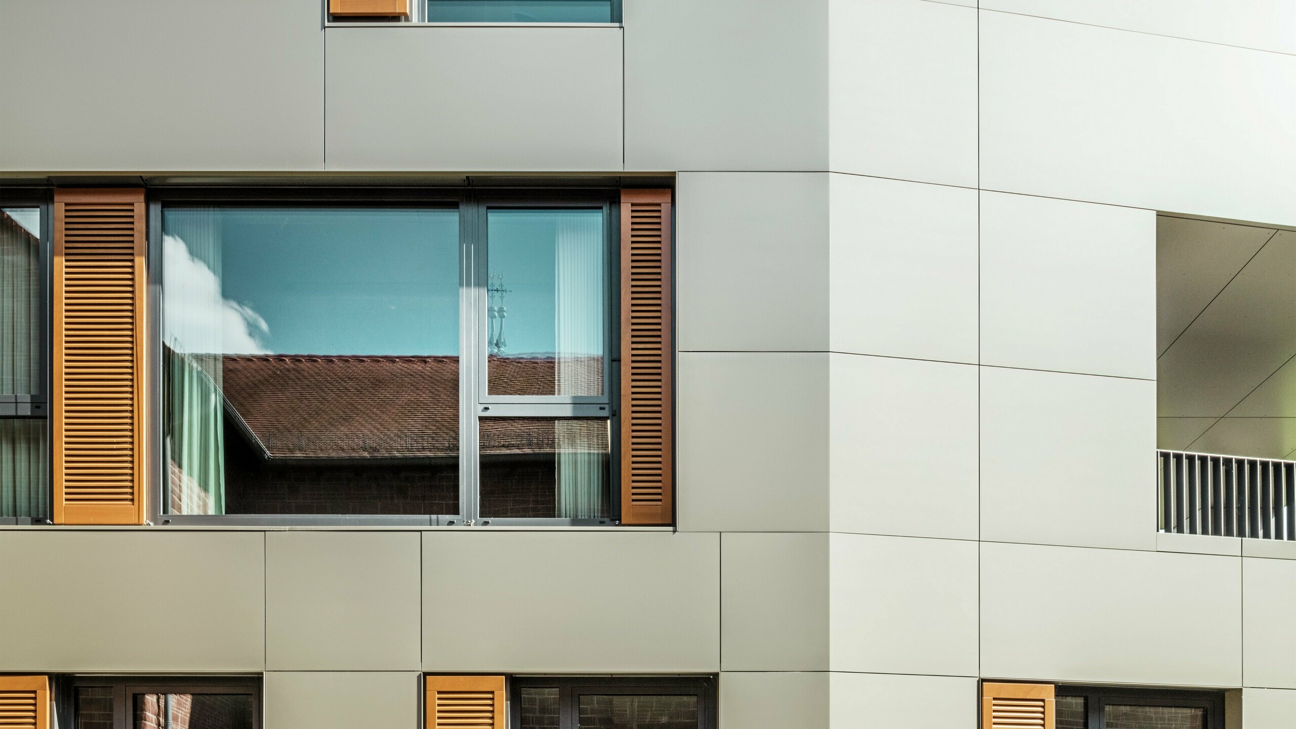 Detailaufnahme der Fassade des Rathauses in Alzenau, Deutschland, mit Aluminium-Verbundplatten von PREFA in der Farbe bronze. Die großen Fenster sind von eleganten Holzjalousien in warmem Braun umrahmt, die der Fassade eine ansprechende Textur verleihen. Im Vordergrund ist eine gepflegte Blumenrabatte zu sehen, die das urbane Design des Gebäudes mit grünen Akzenten ergänzt. Der klare Himmel und die saubere Linienführung der Architektur unterstreichen den modernen Charakter des Gebäudes.