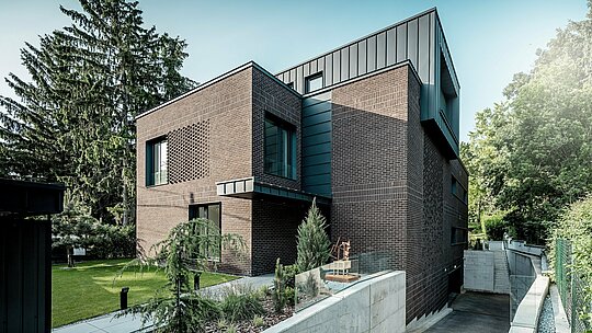 Die Aufnahme zeigt die moderne Architektur der Budapester Mehrgenerationen‑Villa mit ihrer markanten Kombination aus dunkelrotem Klinker und PREFALZ Aluminium in P.10 Anthrazit. Der klar definierte Kubus mit vertikalen Winkelstehfalzen setzt einen starken Kontrast zur strukturierten Backsteinfassade darunter. Die elegant geführte Zufahrt zur Tiefgarage und die sorgfältig gestaltete Gartenanlage betonen die hochwertige Umsetzung. Die langlebigen, witterungsbeständigen PREFA Aluminiumprodukte verleihen dem Gebäude ein zeitloses, präzises und architektonisch ausdrucksstarkes Erscheinungsbild.