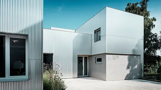 Das Envirocenter Nitra besticht durch seine silbern schimmernde PREFA Aluminiumfassade aus Strangpressprofil Welle in Naturblank. Die vertikalen Wellenstrukturen betonen die klaren, kubischen Baukörper, während präzise eingefasste Fenster und lebendige Schattenspiele spannende Akzente setzen. Die langlebigen, witterungsbeständigen Aluminiumprofile schaffen einen eleganten Kontrast zu den hellen Putzflächen und der naturnahen Bepflanzung.