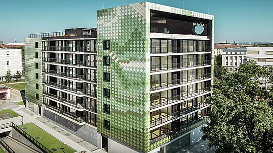 Das Wohn- und Geschäftsgebäude „Pick-Nick“ besticht durch seine markante PREFA-Fassade aus Aluminiumrauten in verschiedenen Grün- und Beigetönen. Das dynamische Farbkonzept und die klare Linienführung der Aluminiumfassade harmonieren mit den großen Glasflächen und den umliegenden Grünanlagen. Der Schriftzug „Pick-Nick“ am Dachrand unterstreicht die Bedeutung des Gebäudes für die Stadt Dresden.