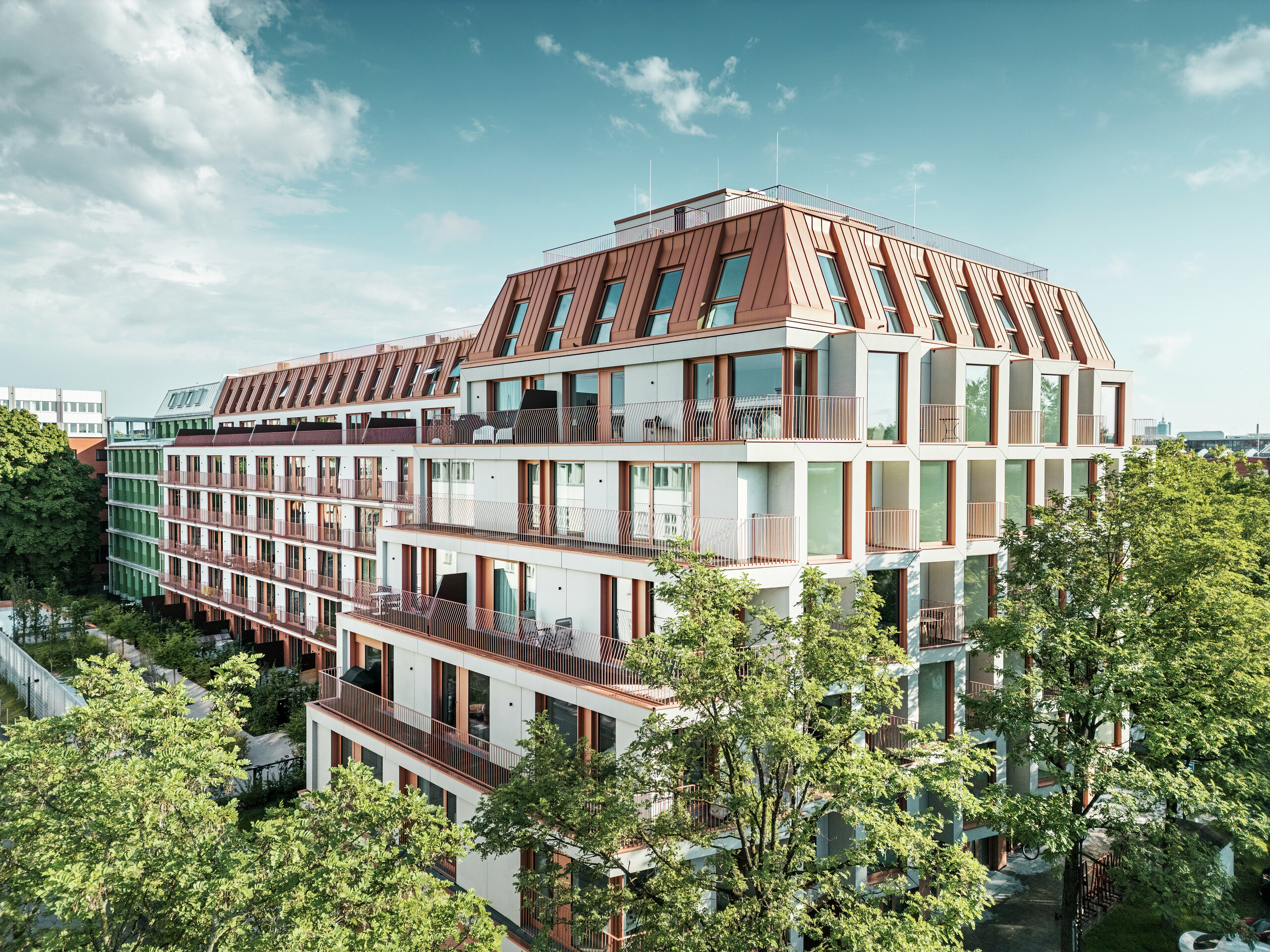 Gesamtansicht des urbanen Wohnprojektes Van B in München mit klarer Architektur, begrünten Außenbereichen und dem markanten PREFALZ Aluminiumdach in IGP Orange HDP von PREFA. Das widerstandsfähige und wartungsarme Dachmaterial bildet das verbindende Gestaltungselement des Gebäudes und sorgt für eine langlebige, ästhetische Lösung im urbanen Raum. Großzügige Fensterflächen, Balkone und durchdachte Grundrisse bieten modernen Wohnkomfort mit hoher gestalterischer Qualität.