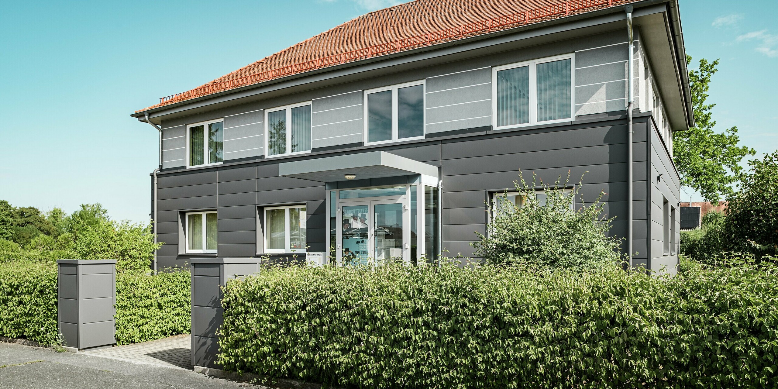 Die modernisierte Fassade der Kaminkehrer-Innung in Bindlach ist mit PREFA Sidings in der Farbe P.10 Dunkelgrau verkleidet. Die Aluminiumpaneele verleihen der frisch sanierten Immobilie eine elegante und zeitgemäße Ausstrahlung. Die präzise verlegten Paneele, die bis zum Eingang führen, schützen das Gebäude effektiv und sorgen für eine nachhaltige Fassadengestaltung. Das Material ist pflegeleicht, langlebig und fügt sich harmonisch in die natürliche Umgebung ein.