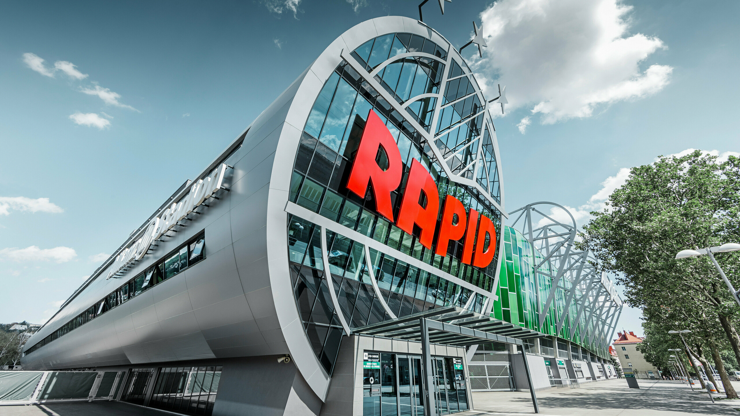 Ansicht des Allianz Stadions von SK Rapid Wien mit auffälliger PREFA Fassade in Form der Aluminium Verbundplatte in Silbermetallic. Die Gebäudehülle strahlt Dynamik und Moderne aus. Der Eingangsbereich, gekrönt mit dem leuchtend roten RAPID-Schriftzug, lädt ein in das Herz des Fußballs, eingefasst von einer futuristischen und zugleich eleganten Fassadenstruktur, die Sportarchitektur auf ein neues Niveau hebt.