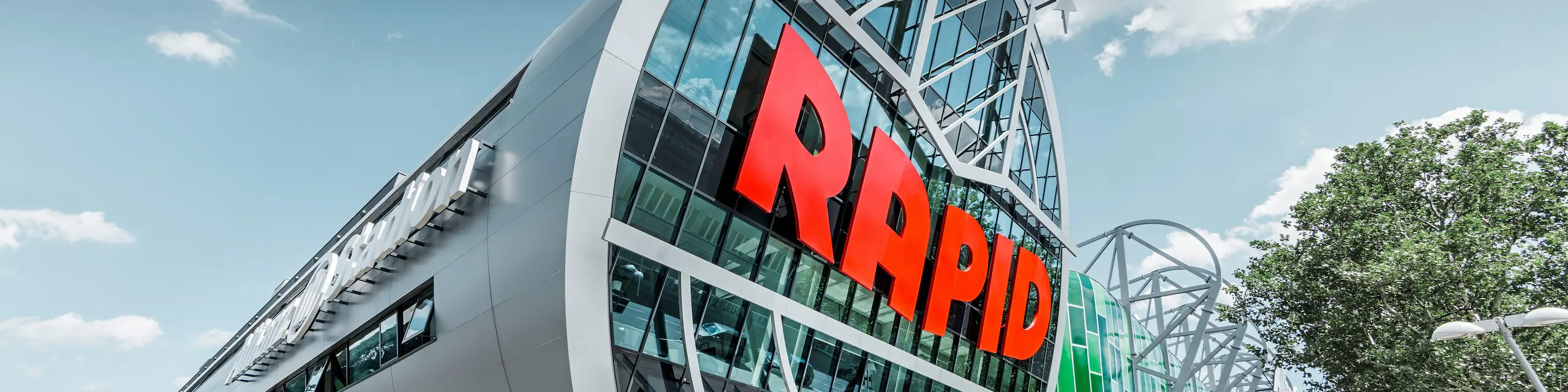 Ansicht des Allianz Stadions von SK Rapid Wien mit auffälliger PREFA Fassade in Form der Aluminium Verbundplatte in Silbermetallic. Die Gebäudehülle strahlt Dynamik und Moderne aus. Der Eingangsbereich, gekrönt mit dem leuchtend roten RAPID-Schriftzug, lädt ein in das Herz des Fußballs, eingefasst von einer futuristischen und zugleich eleganten Fassadenstruktur, die Sportarchitektur auf ein neues Niveau hebt.