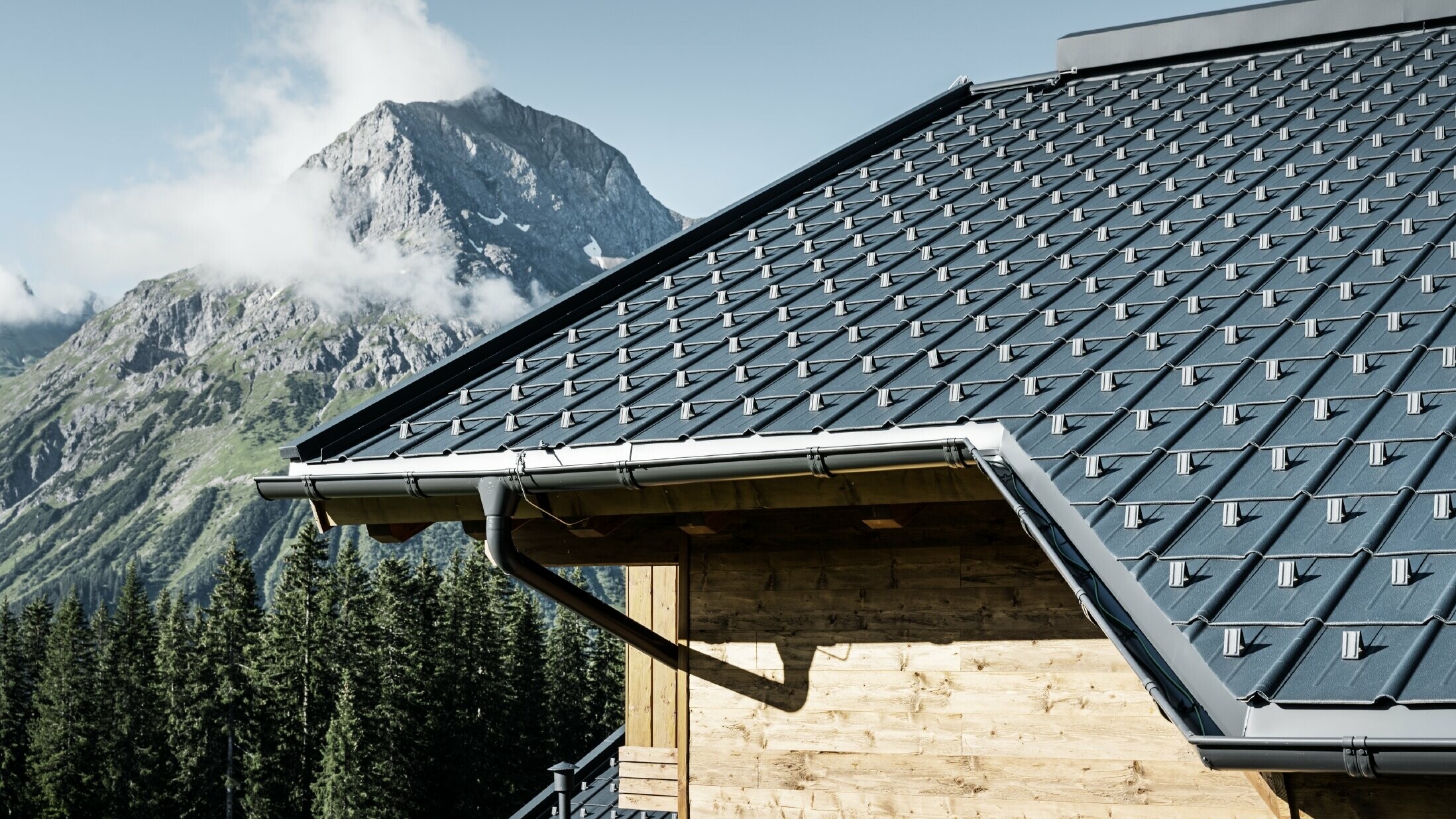 PREFA Dachplatte im alpinen Einsatz - Dachplatte in Anthrazit verlegt auf Hütte in Lech am Arlberg mit Berge im Hintergrund; PREFA Dachrinne in Anthrazit; Holzfassade